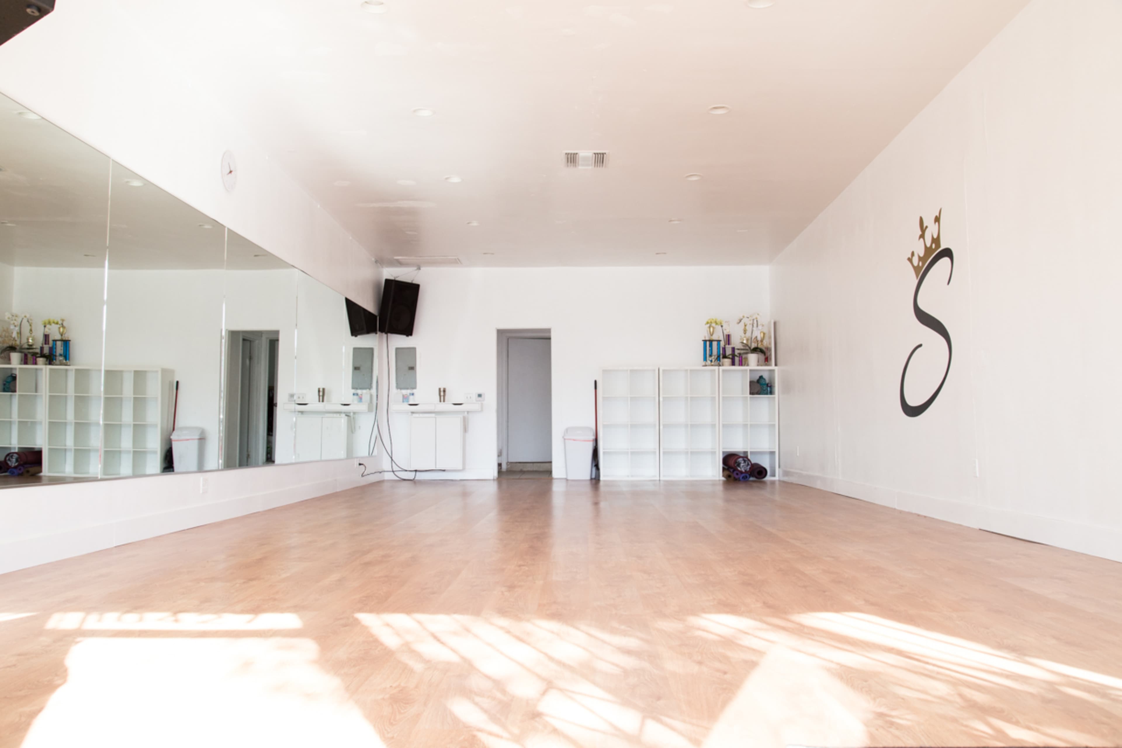 The image shows a spacious dance studio with a mirrored wall, wooden floor, and a large wall-mounted letter "S."