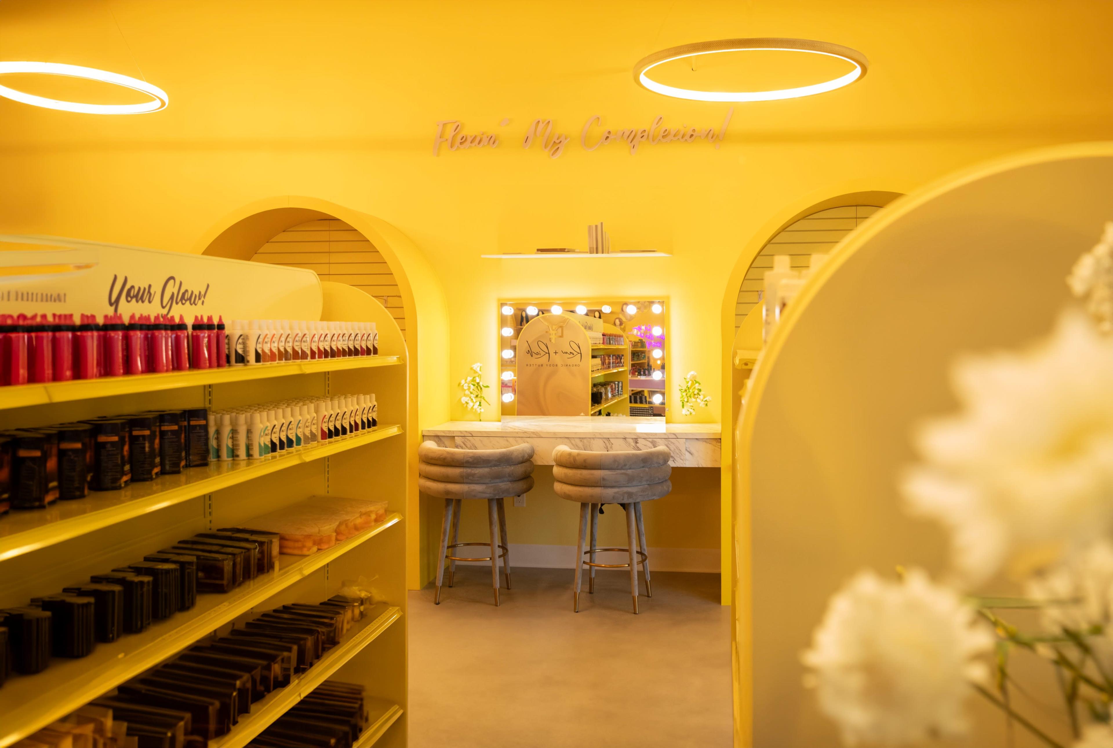 The image shows a brightly colored yellow interior of a beauty store, featuring shelves stocked with products and a vanity area with two stools.