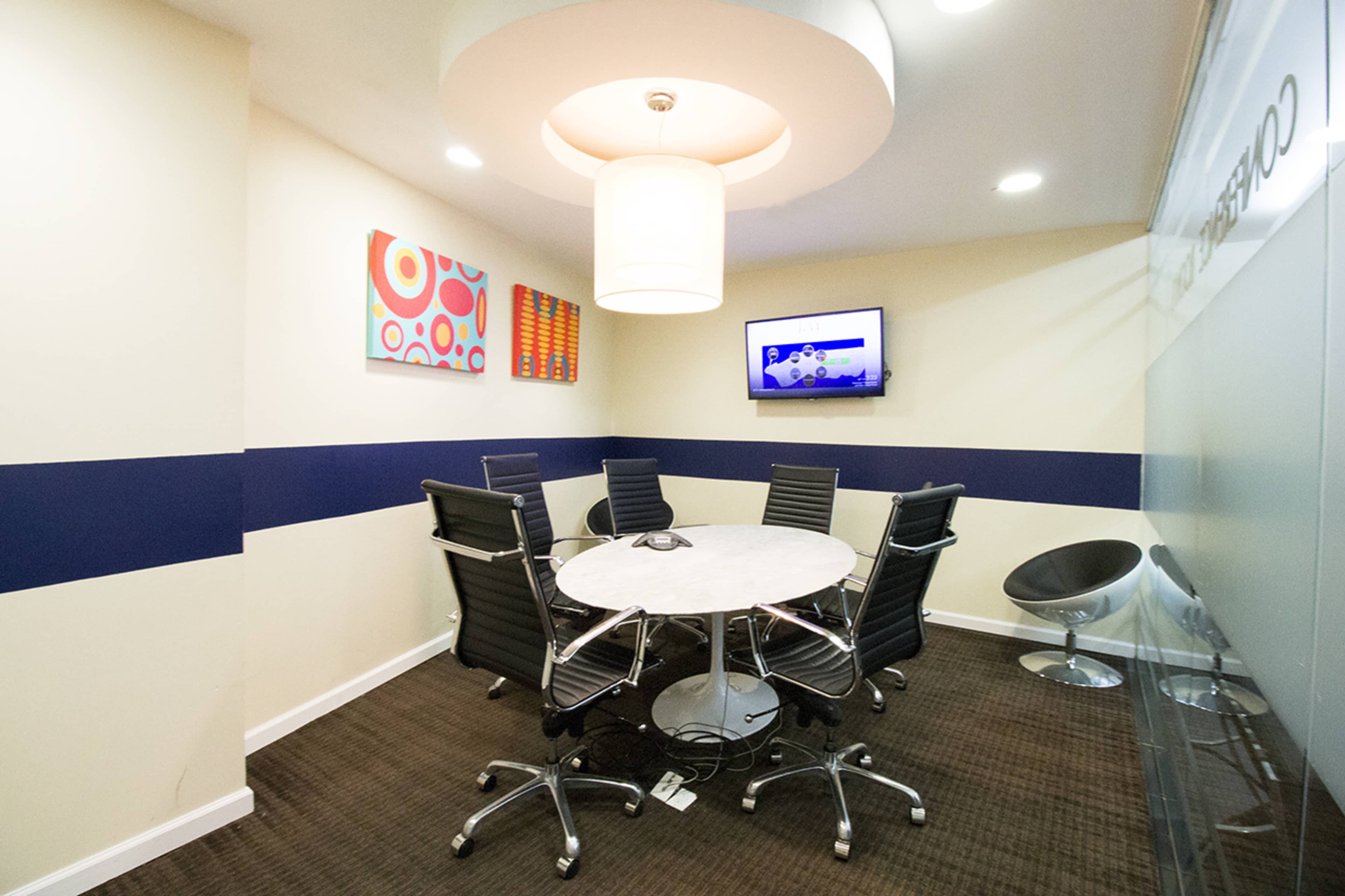 A conference room features a circular table surrounded by six black chairs, with artwork on the walls and a television mounted in the corner.