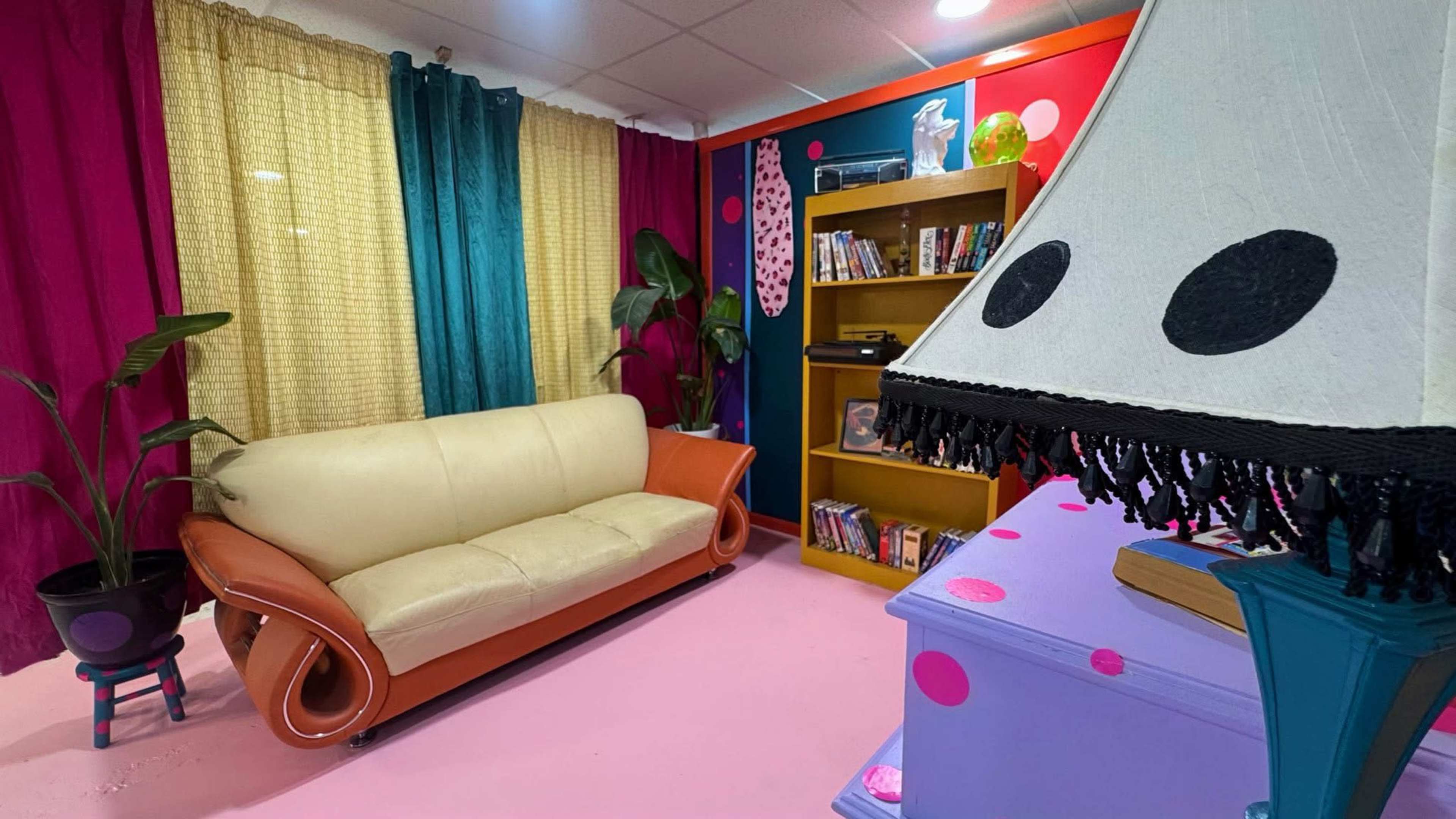 The image shows a colorful living room with a tan and orange sofa, a bookshelf filled with DVDs, and vibrant curtains draping the walls.