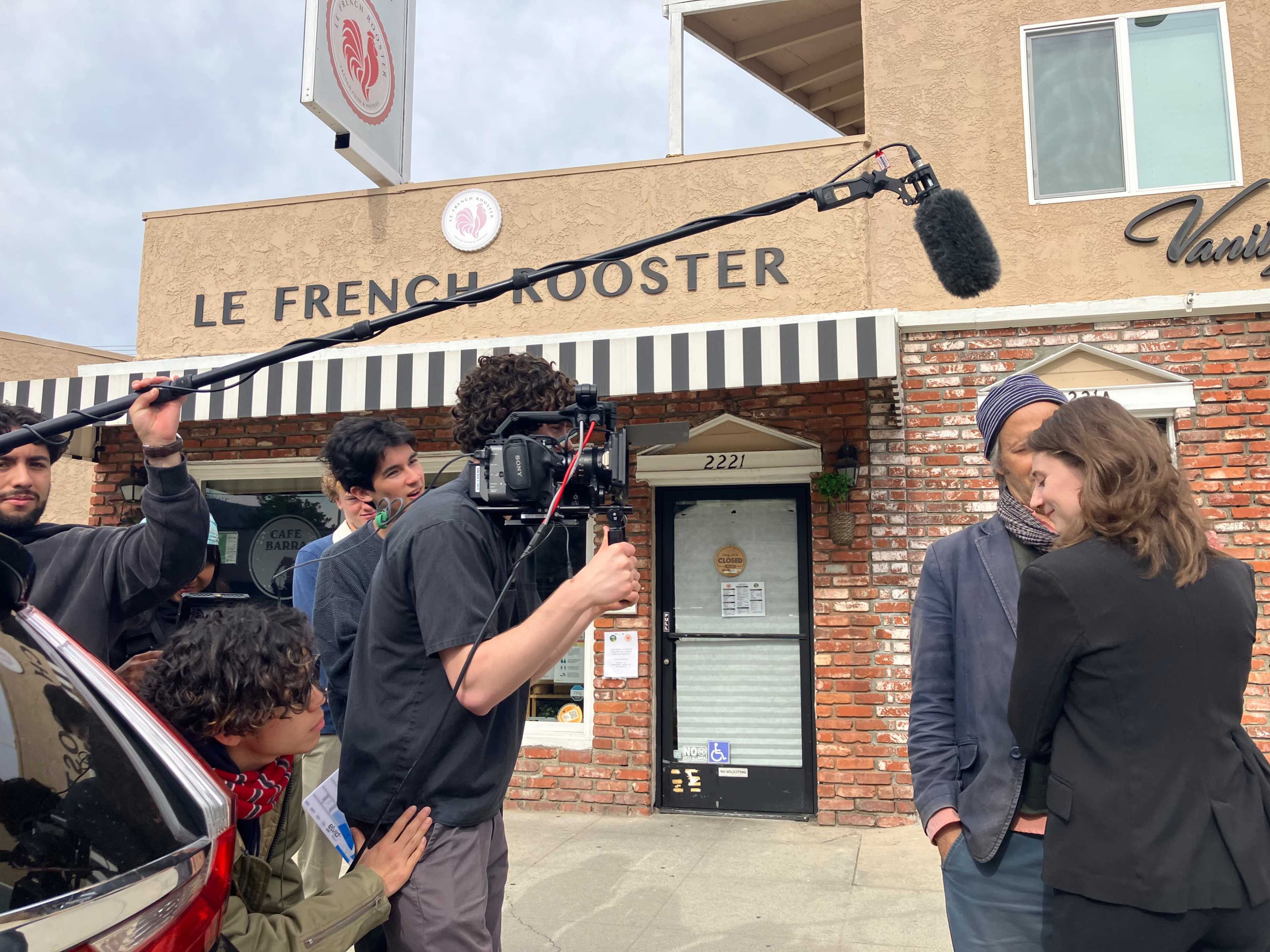 A film crew is recording a scene outside Le French Rooster, with a boom microphone extended over two actors in conversation.