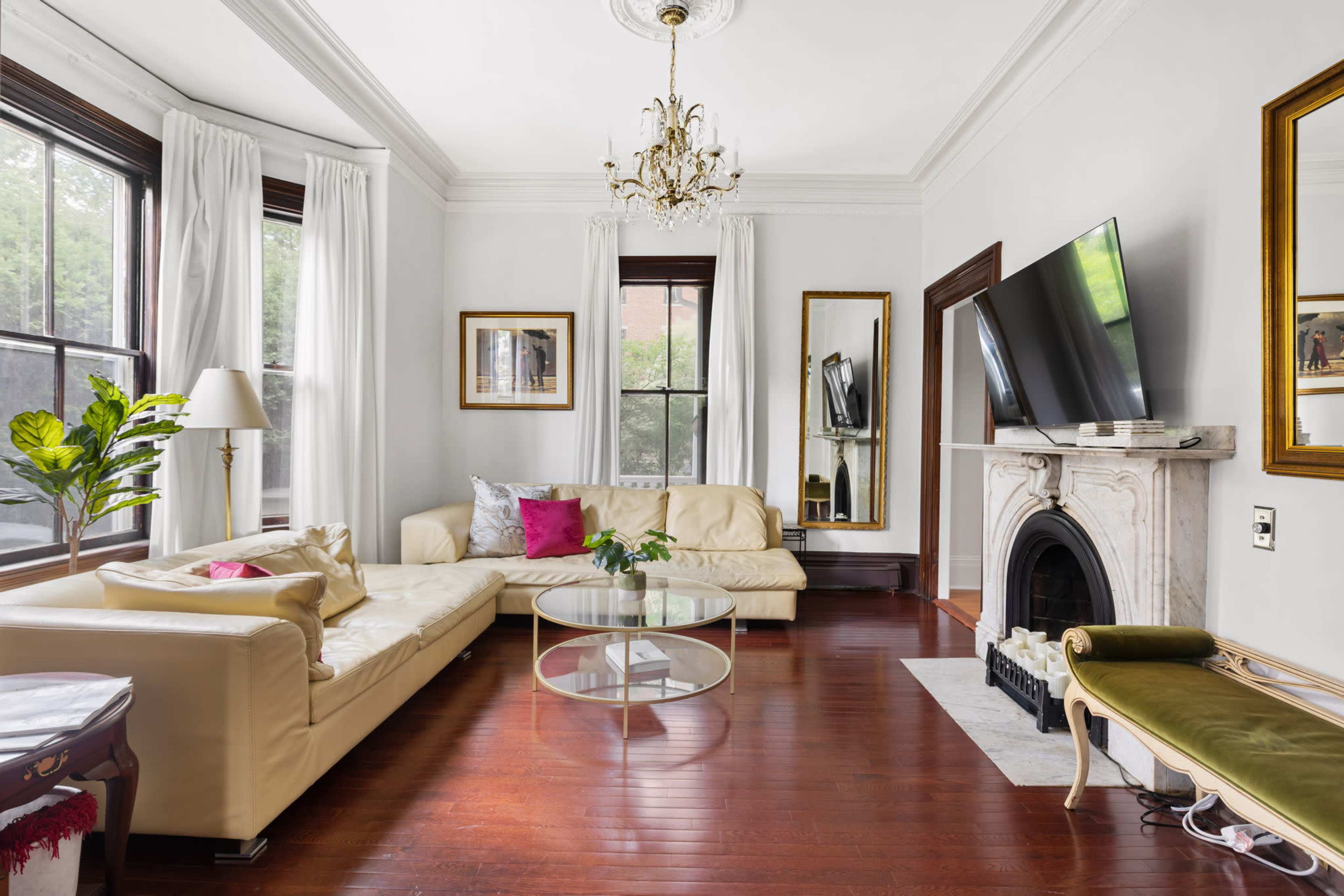 A living room features a cream-colored leather sectional sofa, a glass coffee table, a decorative fireplace, and large windows with white curtains.