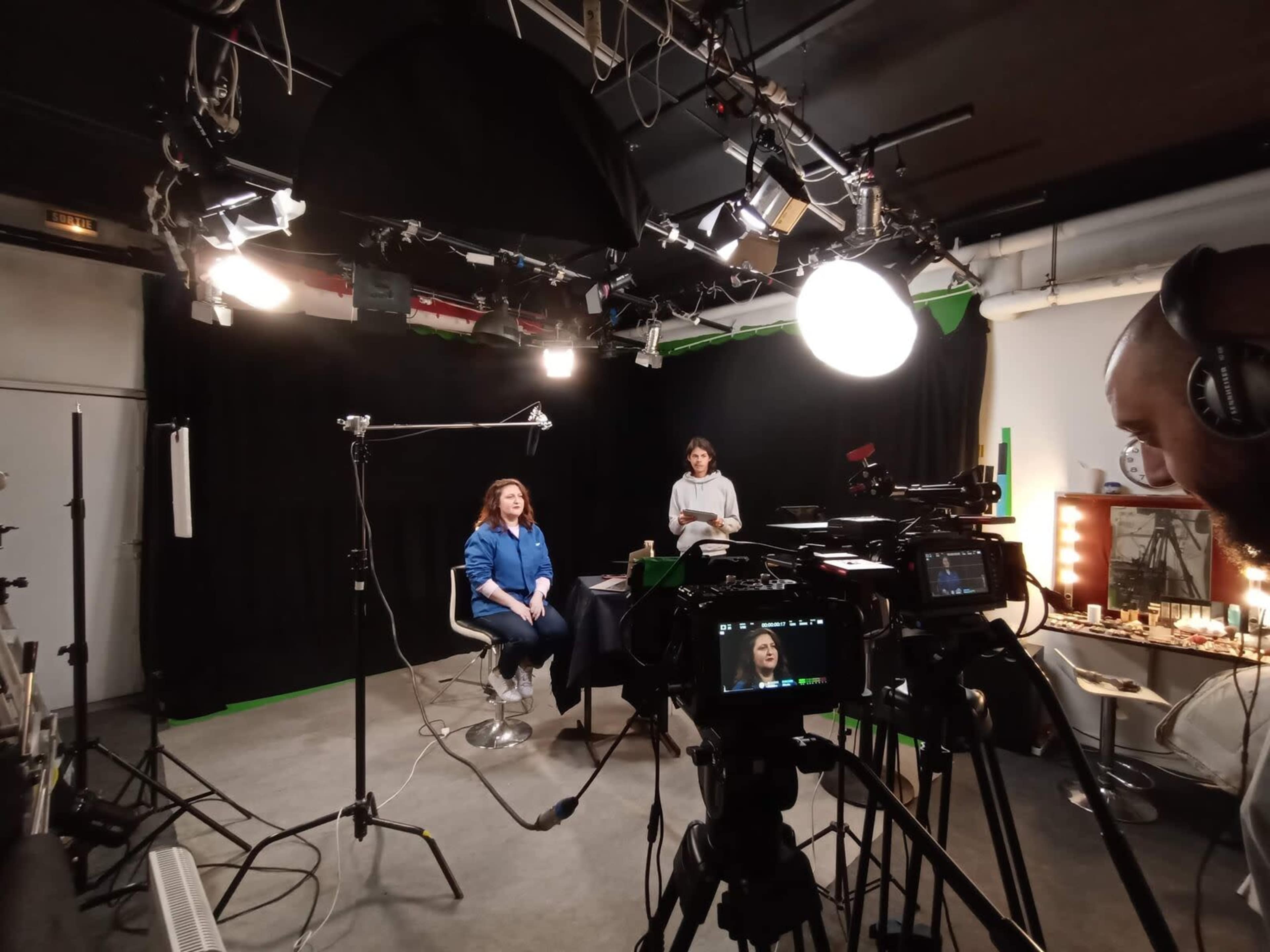 A television studio setup features a woman sitting in a chair under bright lights, while another person operates a camera nearby.