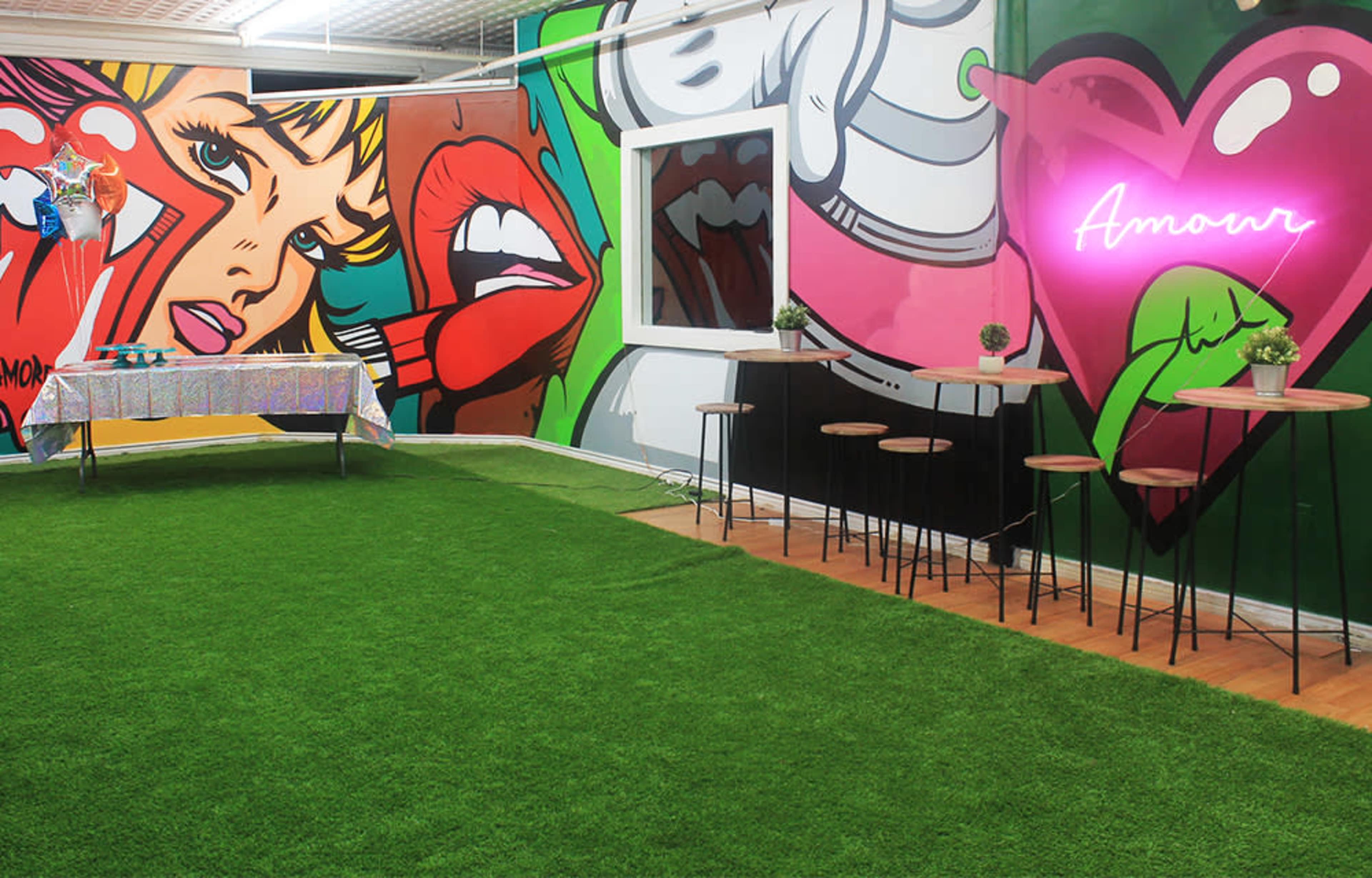 The scene features a colorful interior with vibrant wall art, a grassy floor, a table covered in a shiny cloth, and several high stools arranged around a small seating area.