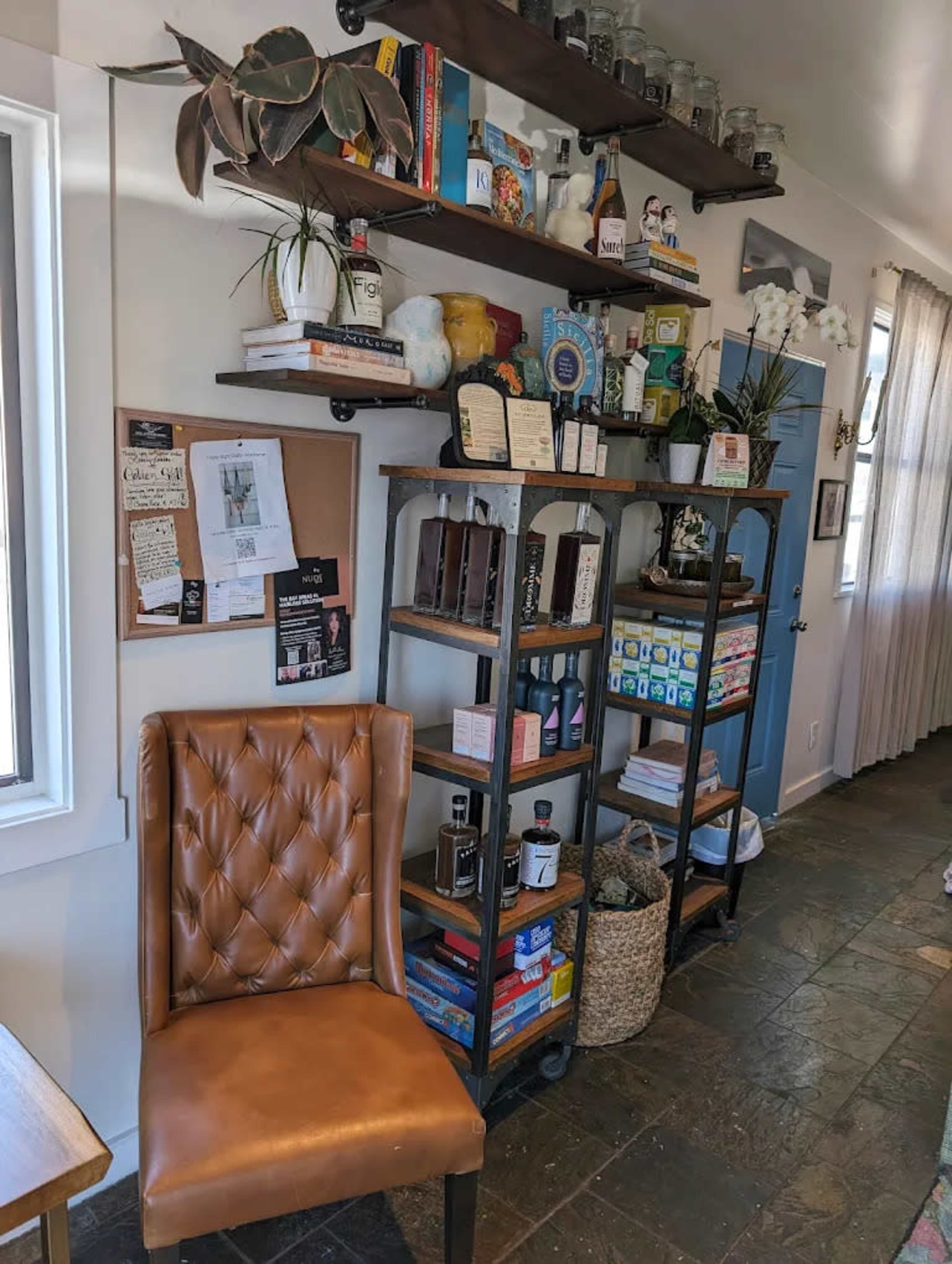 A brown leather chair sits next to a tall shelving unit filled with books, jars, and various decorative items along a wall with a bulletin board and a blue door.