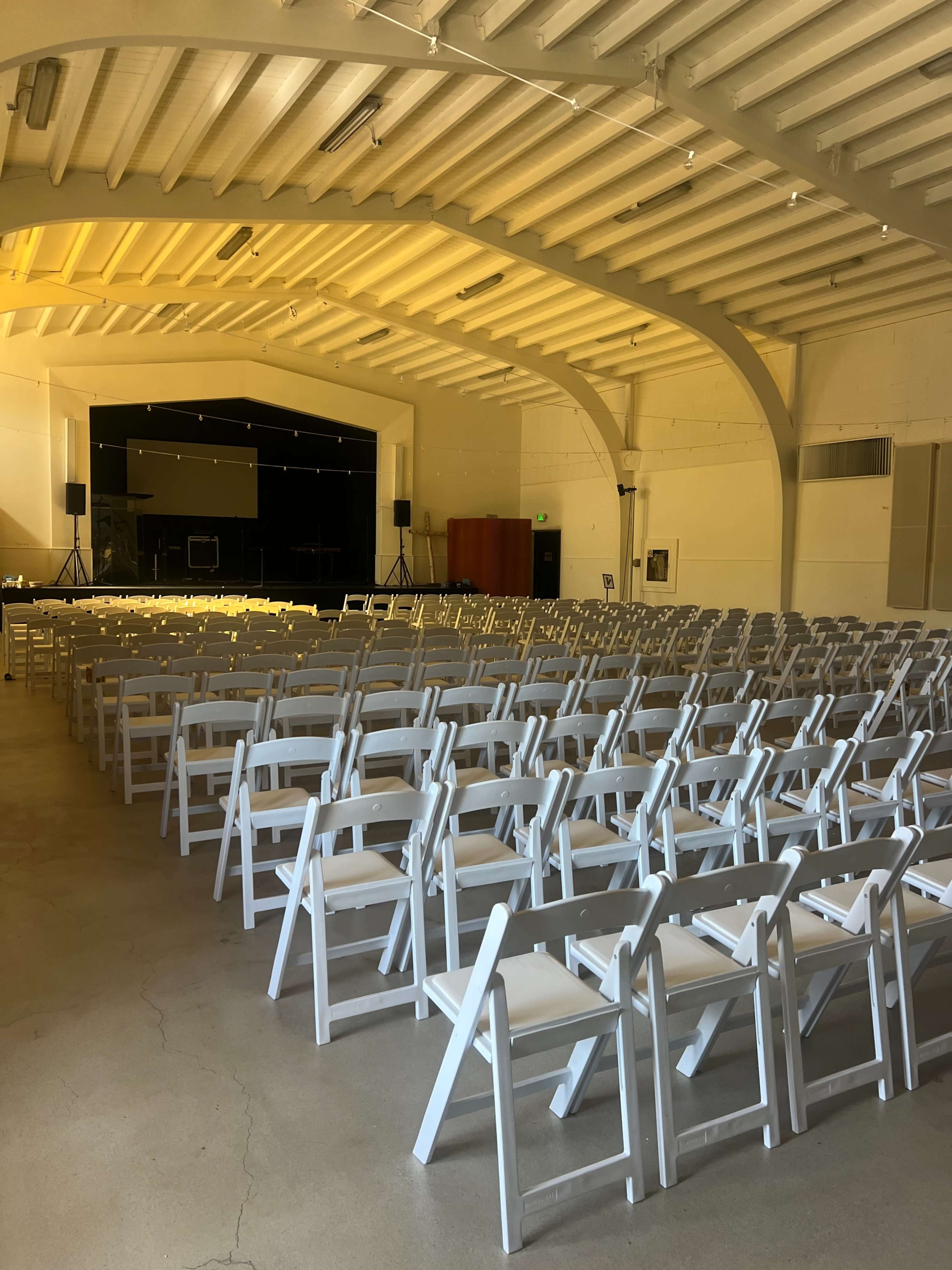 The image shows a large, empty indoor venue with rows of white folding chairs arranged for an event, illuminated by overhead lighting.