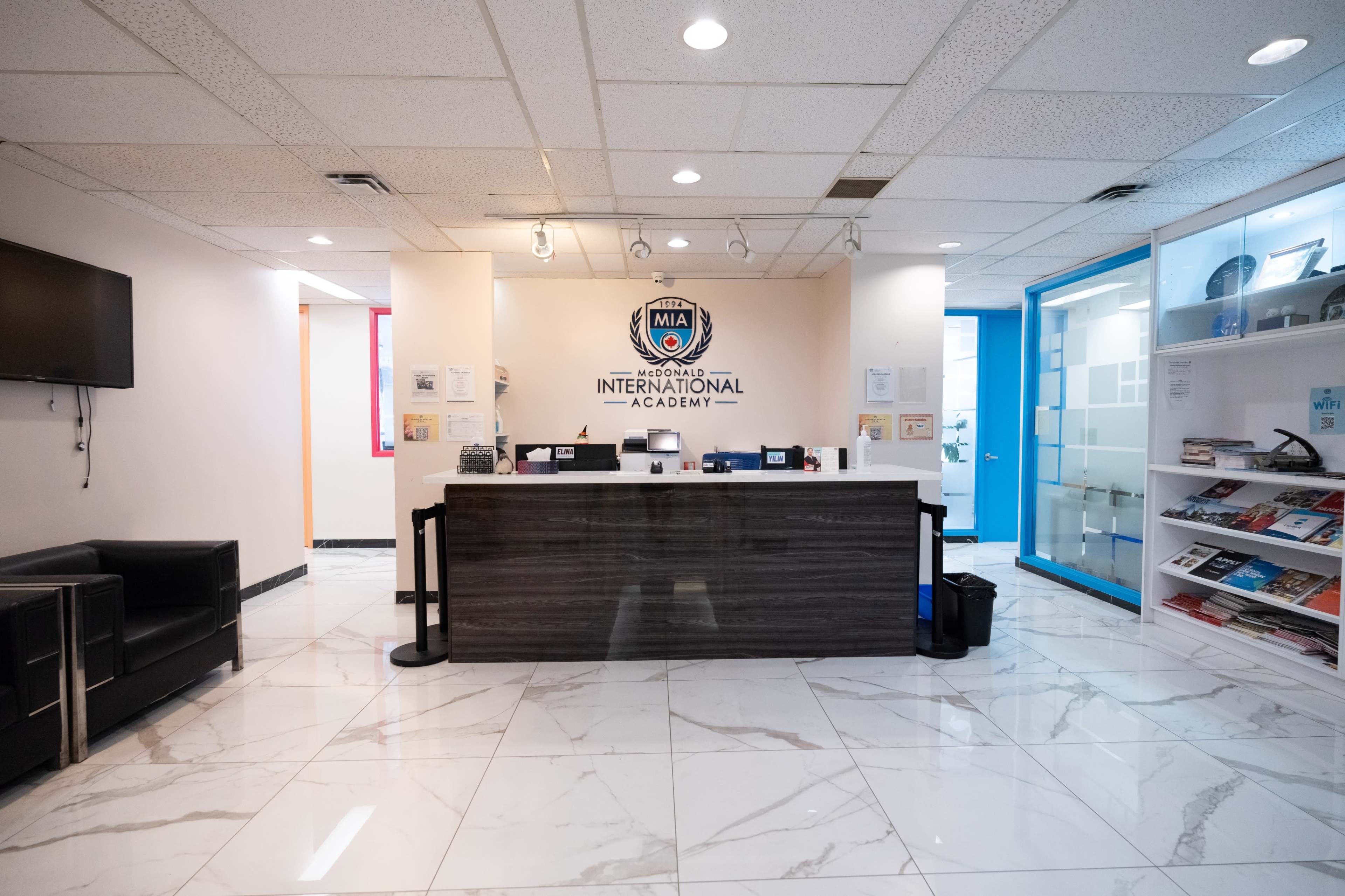 The image shows a reception area of the MIA International Academy, featuring a desk with an academy logo, seating, and informational materials neatly arranged on a shelf.
