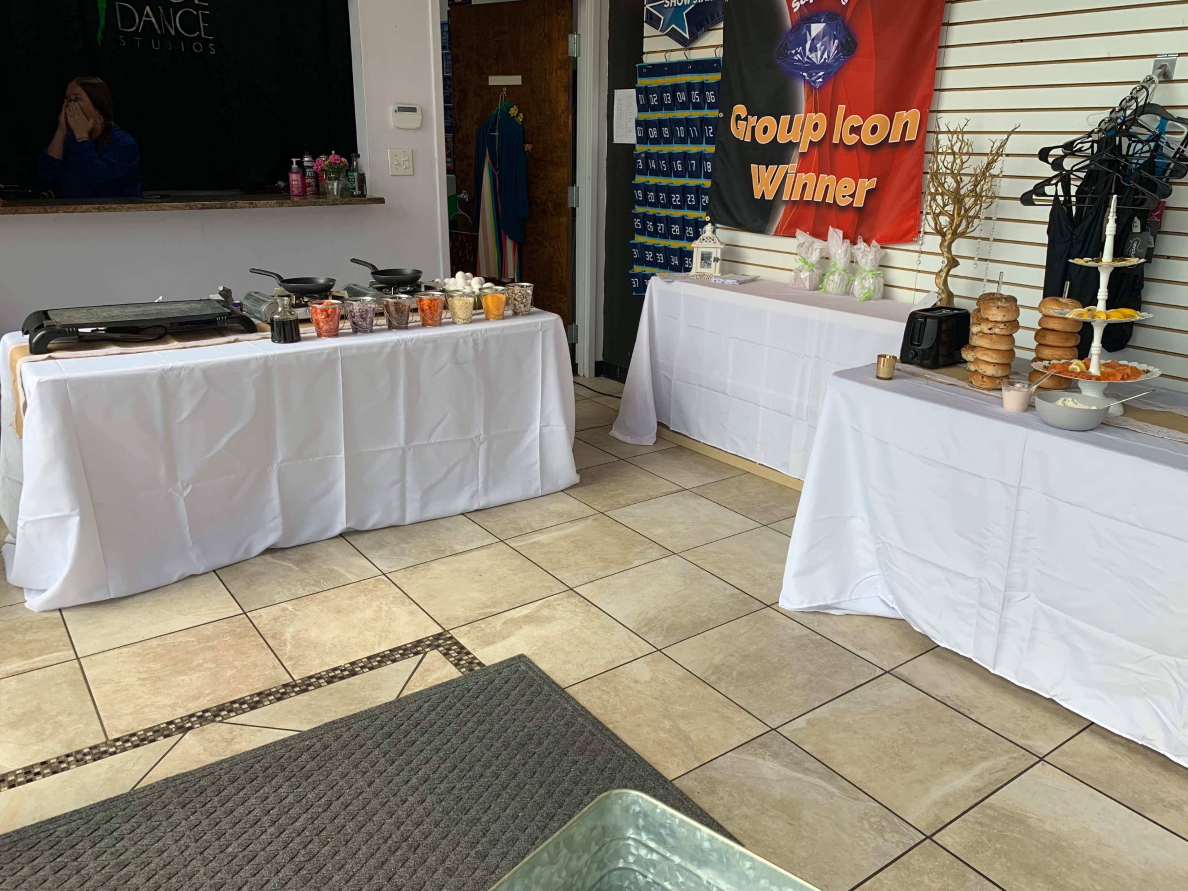 The image shows a room set up for an event, featuring two tables covered with white cloths; one table displays various food items and drinks, while the other holds a tiered display of donuts and an arrangement of snacks.