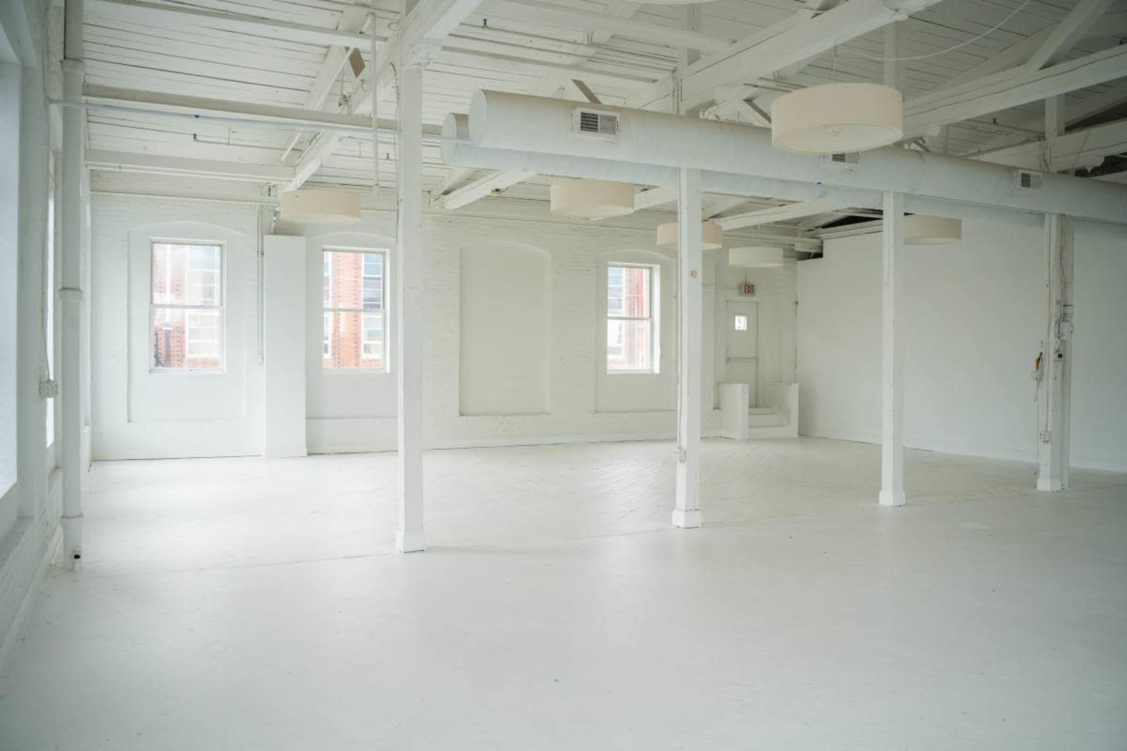 The image shows a spacious, empty room with white walls and ceilings, featuring several windows and cylindrical light fixtures.