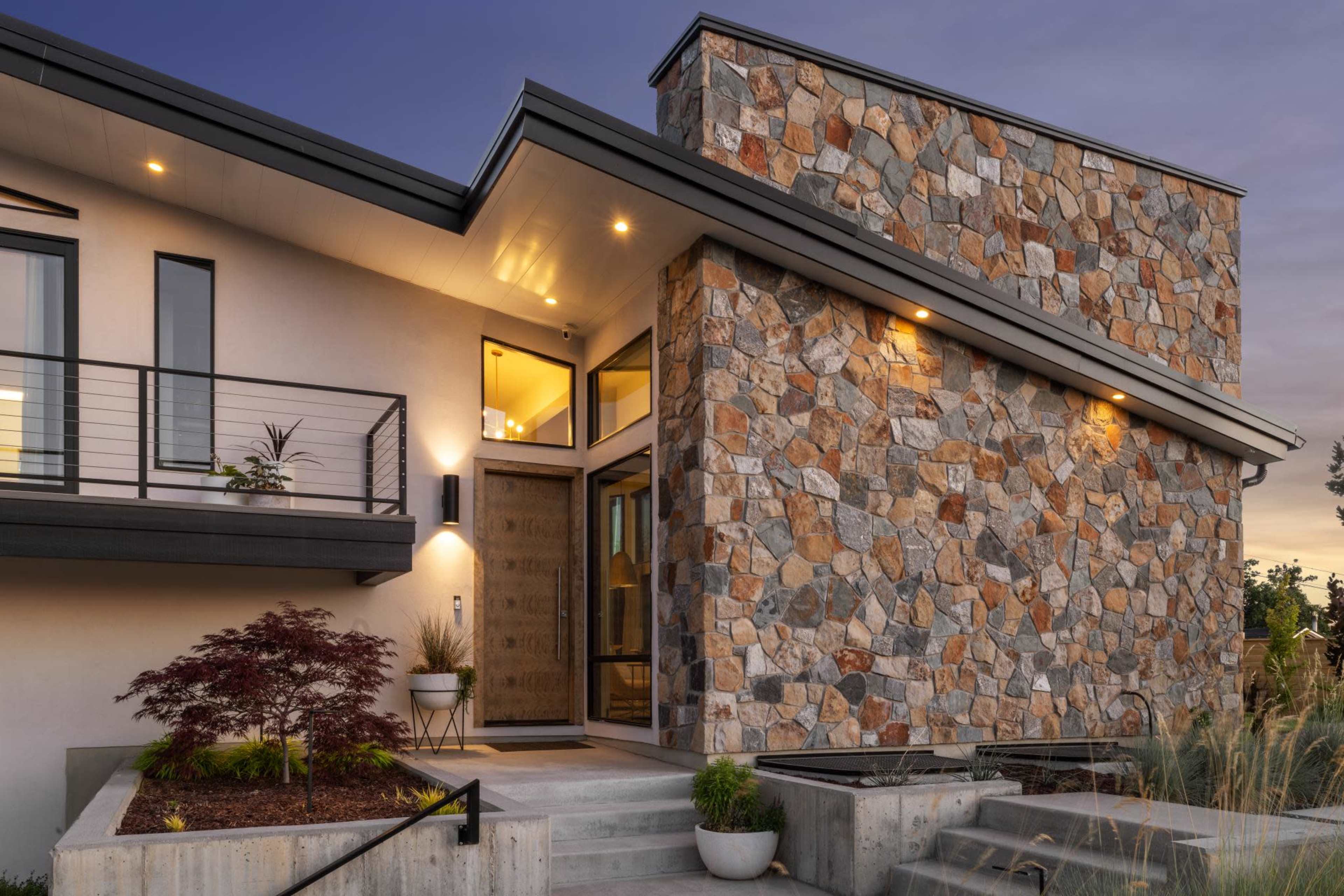 The image shows a modern house with a stone facade, featuring a large entrance with double doors and a landscaped pathway leading to it.