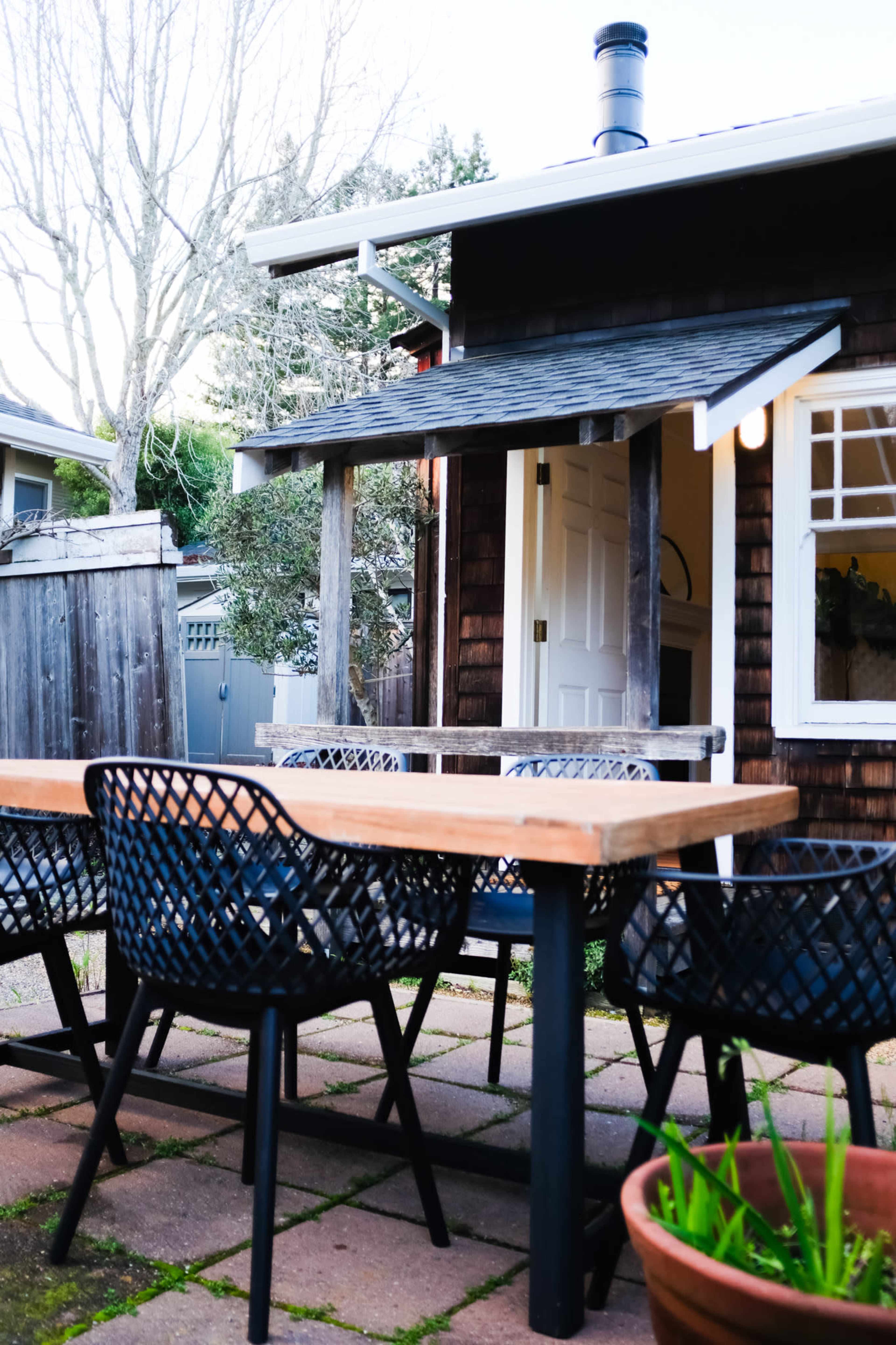 A wooden table with black woven chairs is set in a patio area beside a small house with a chimney and an open door.
