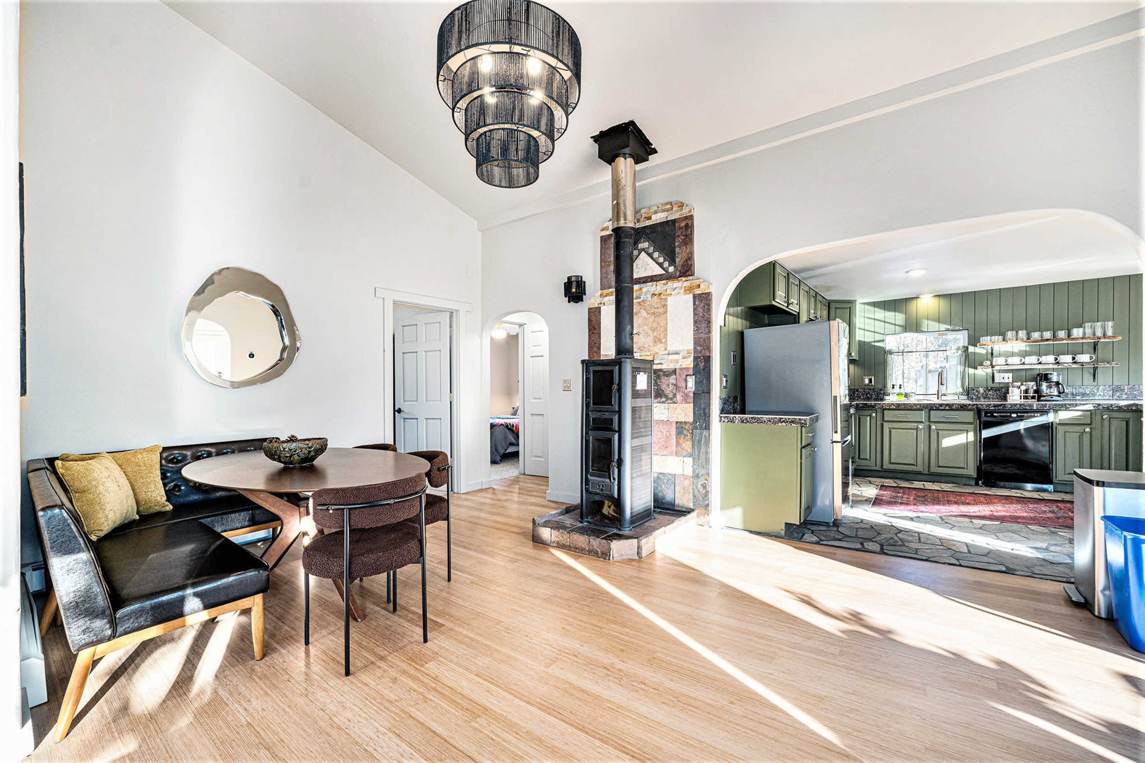 The image shows a modern living space with a round dining table, four chairs, a tall chandelier, and a wood stove, leading into a kitchen area with green cabinetry.