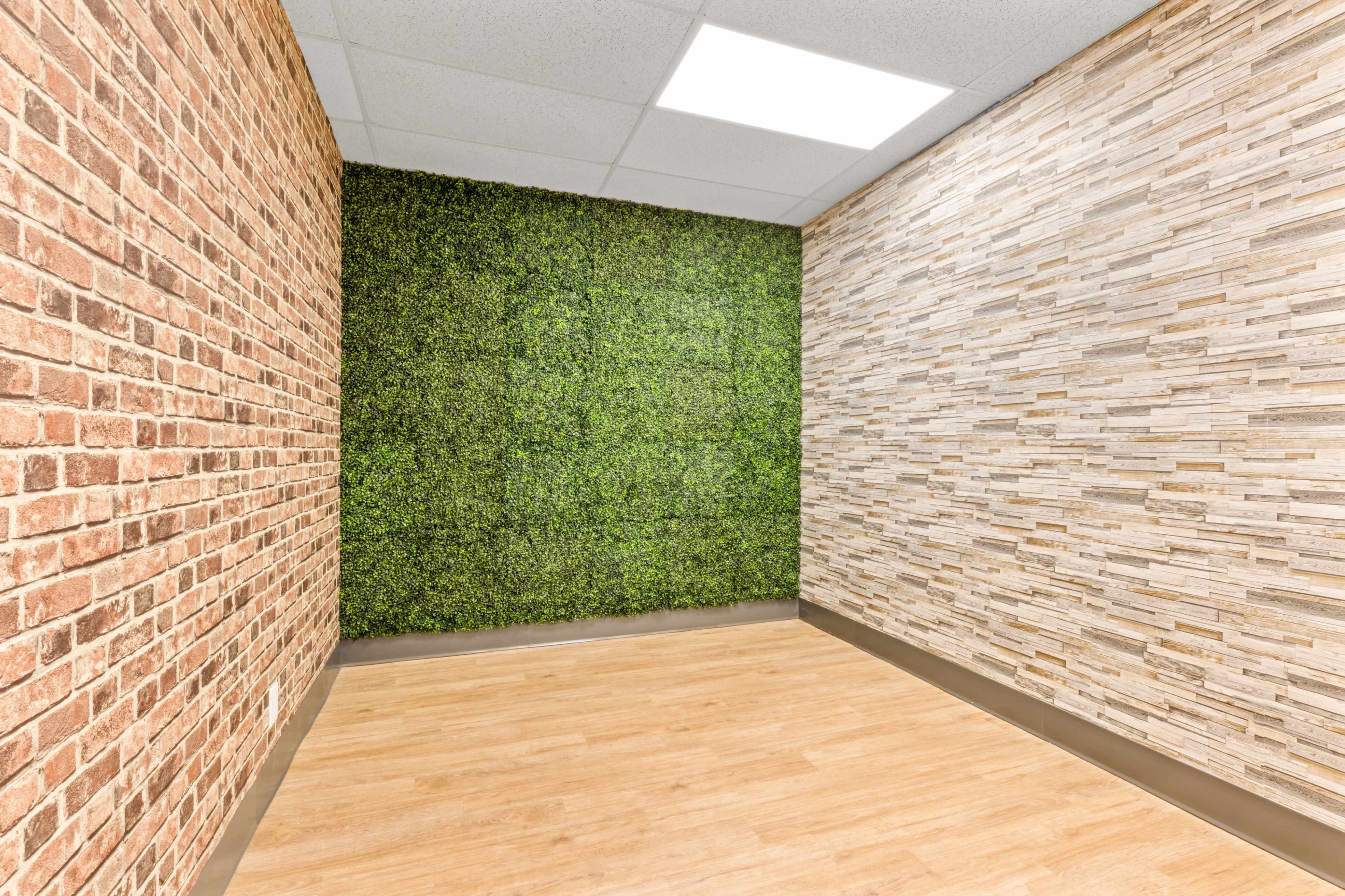 The image shows a corner of a room featuring one wall with exposed brick, another with faux greenery, and a third with stone-like panels, all on a light wood floor.