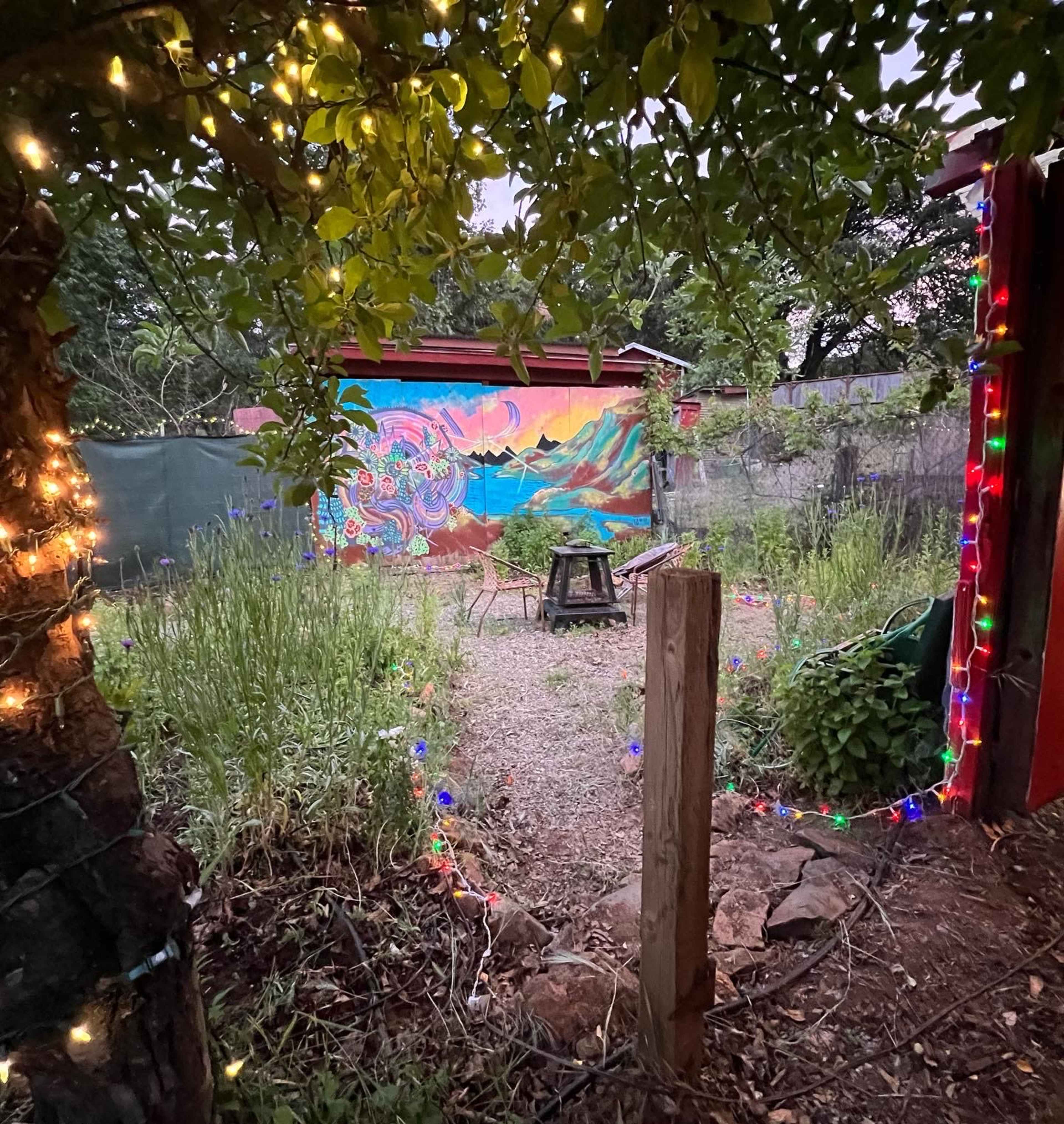 A vibrant mural decorates the back wall of a garden surrounded by plants and lined with colorful string lights.