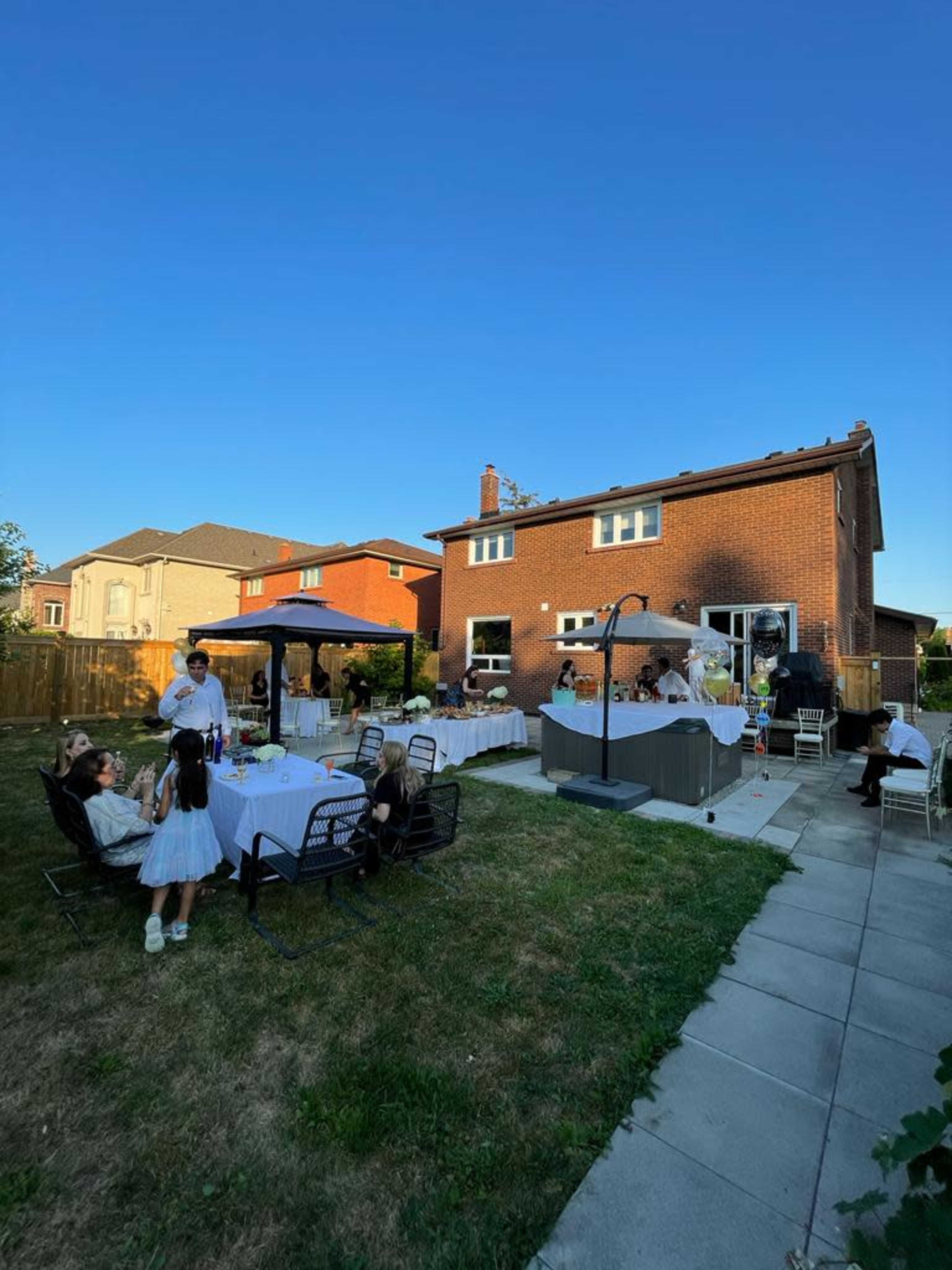 A backyard gathering features tables set for dining, a gazebo, and guests socializing near a house with a clear blue sky above.