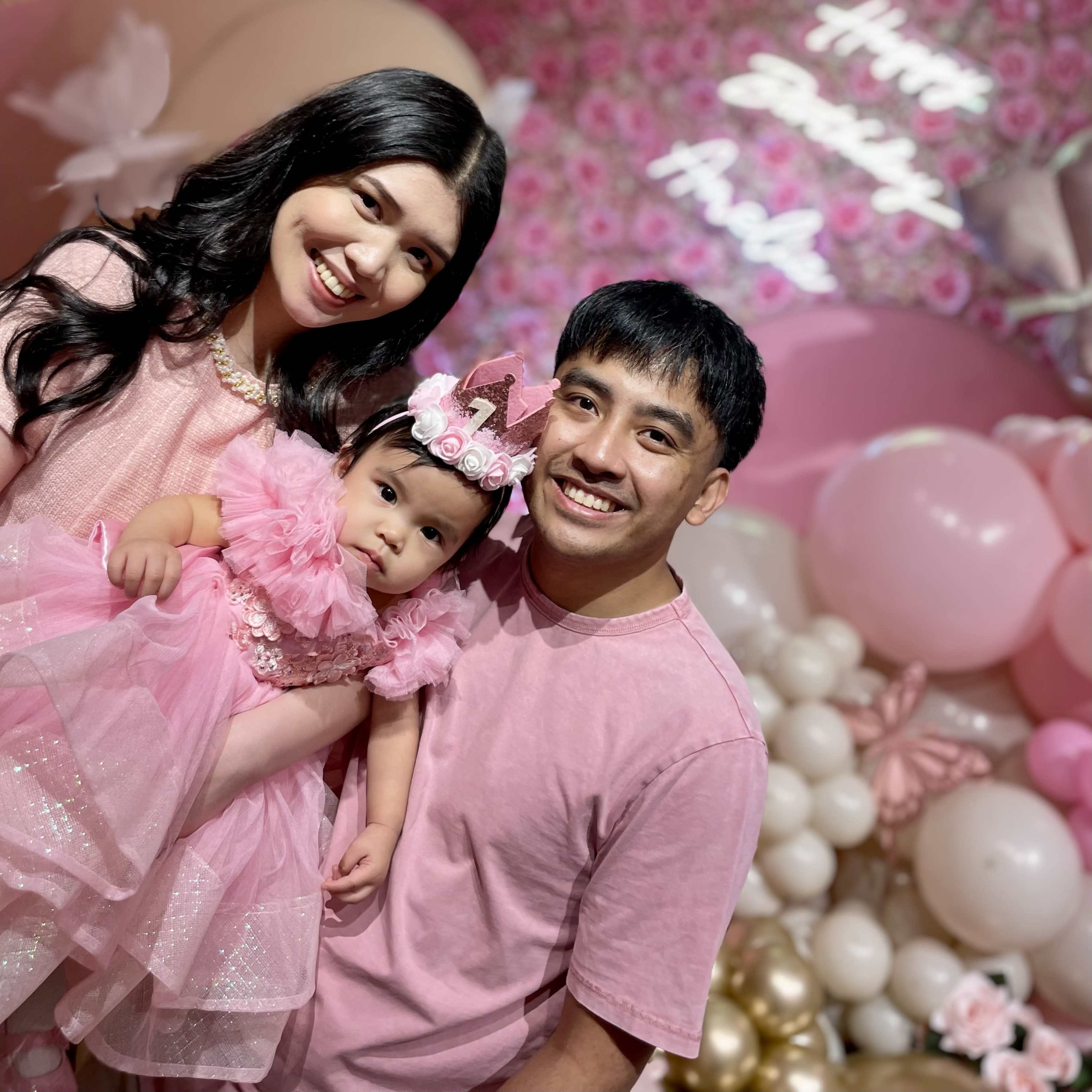 A family poses together for a photo at a birthday celebration, surrounded by pink decorations and balloons.