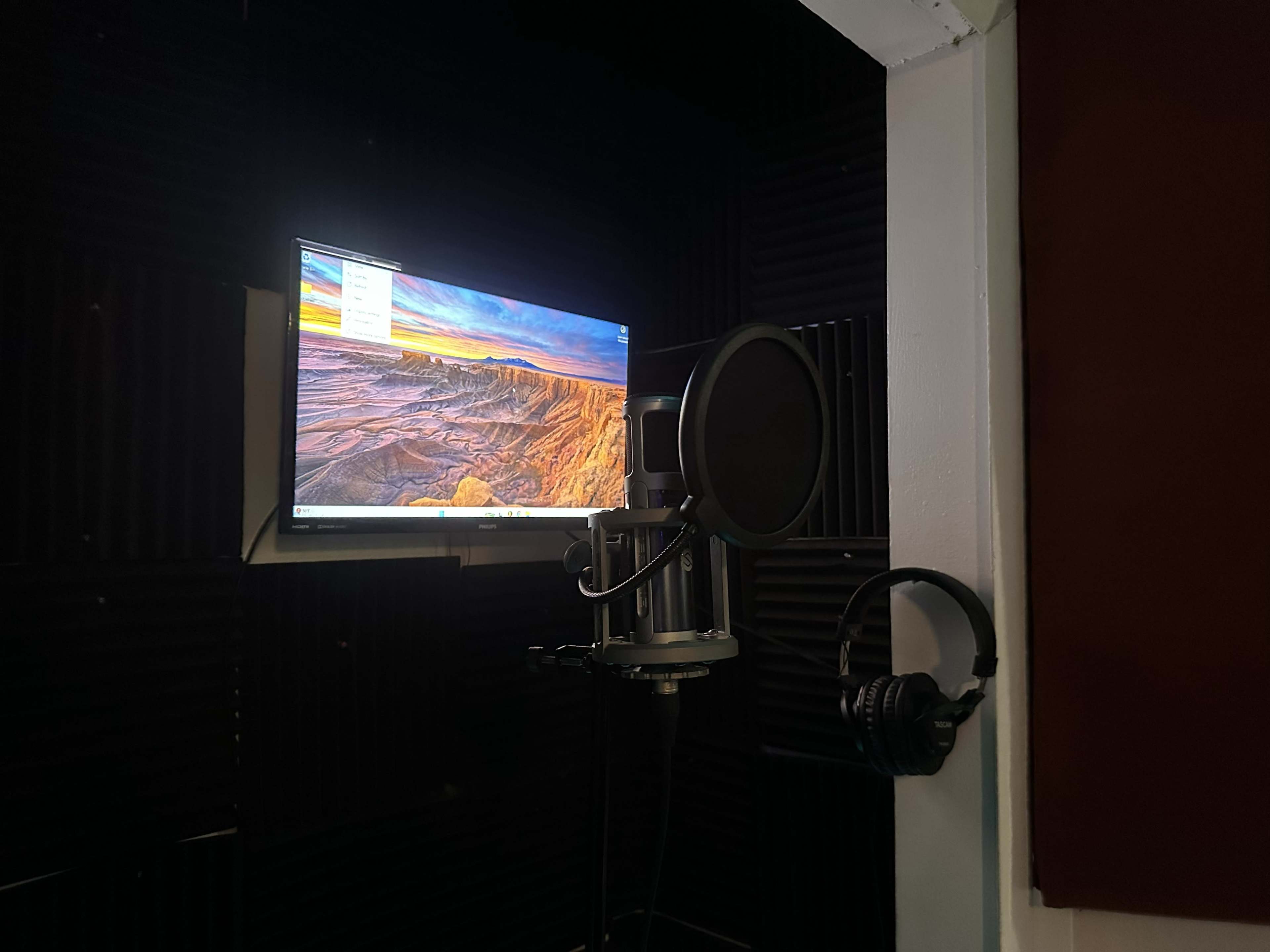 The image shows a small recording booth with a microphone set up next to a mounted computer monitor displaying a landscape image, along with headphones hung on the side.