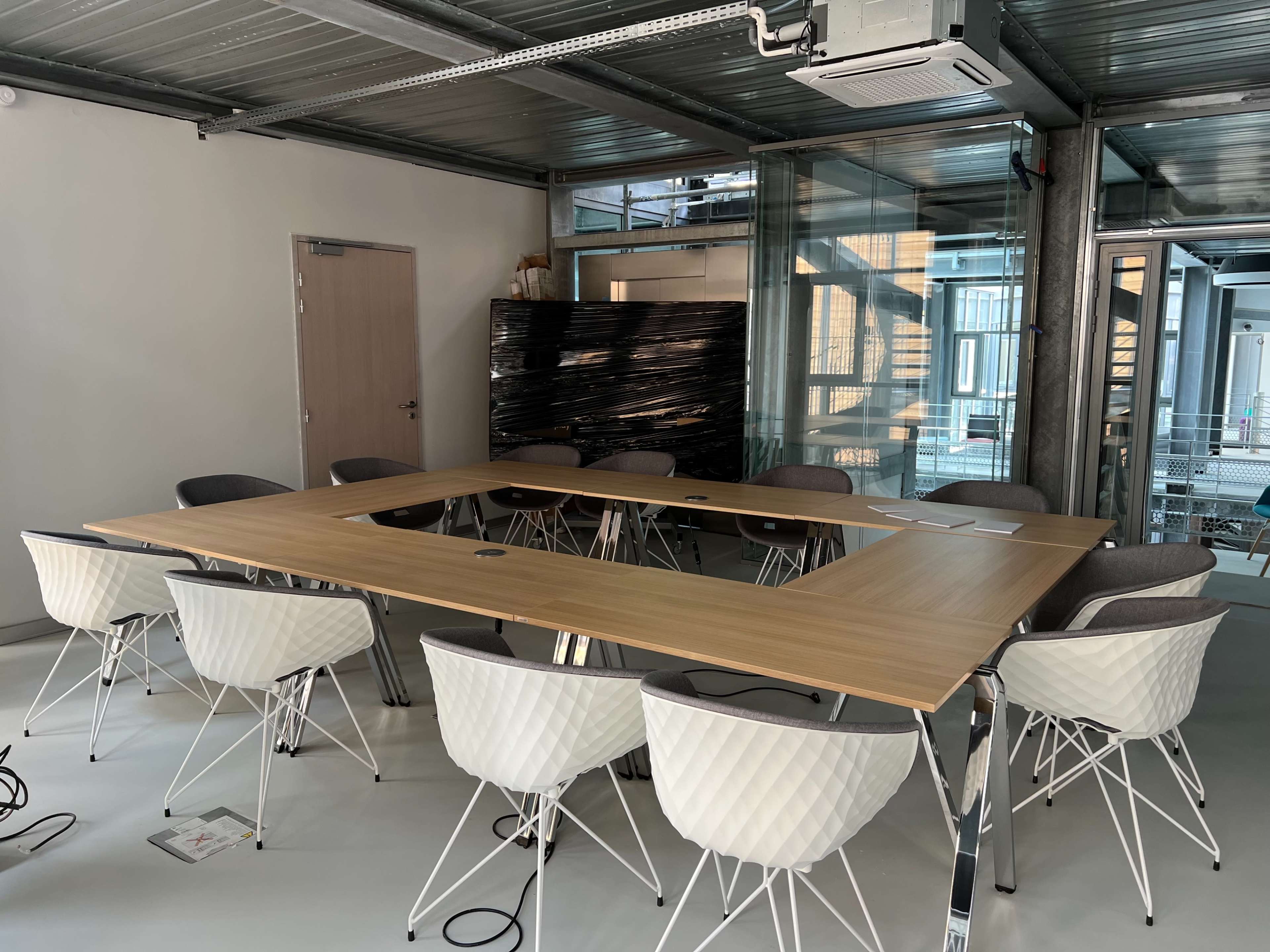 A conference room features a large wooden table surrounded by ten chairs, with glass walls and industrial style elements.