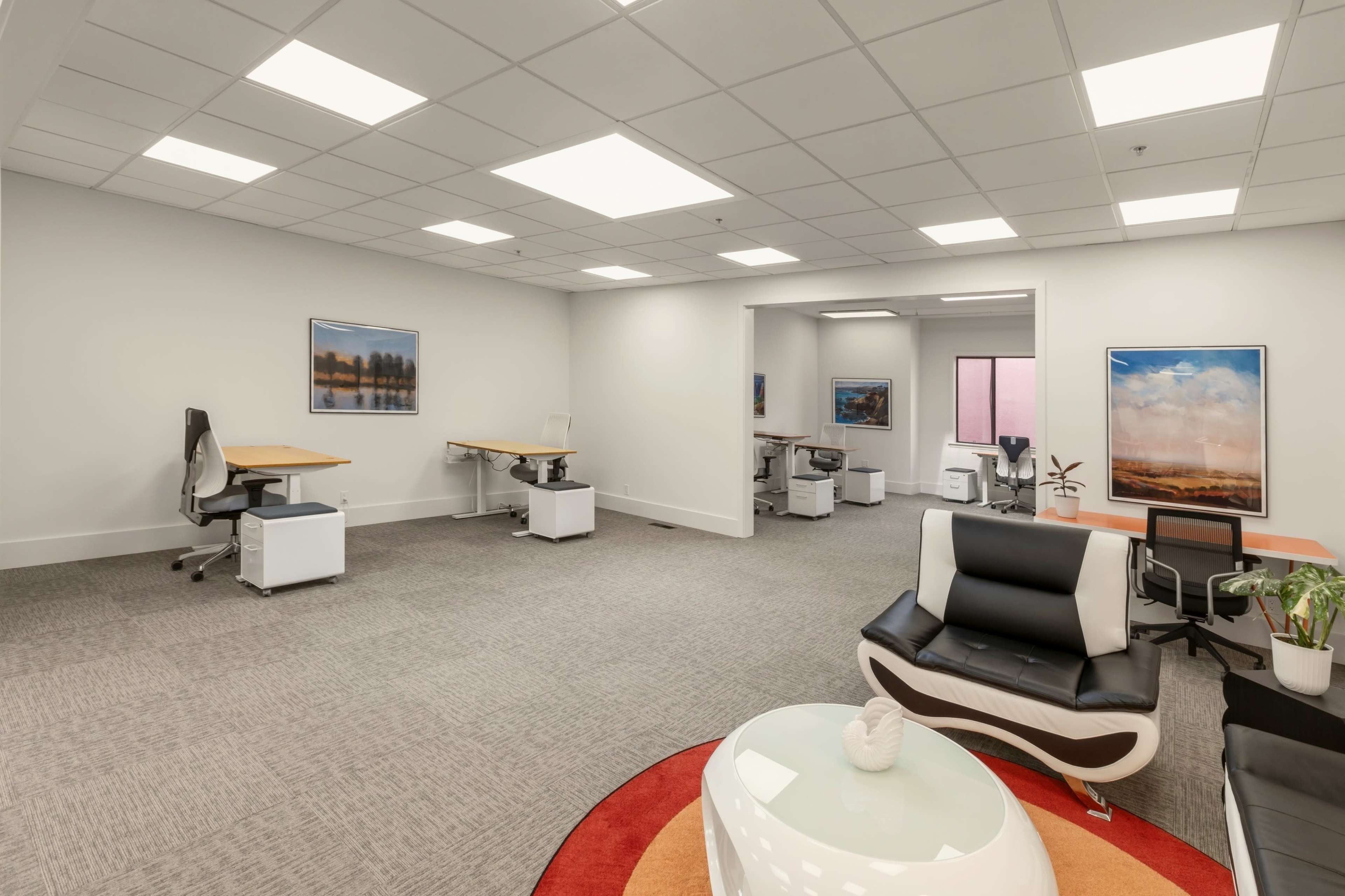 The image shows a modern office space with multiple workstations, a seating area, and a large rug in the center.
