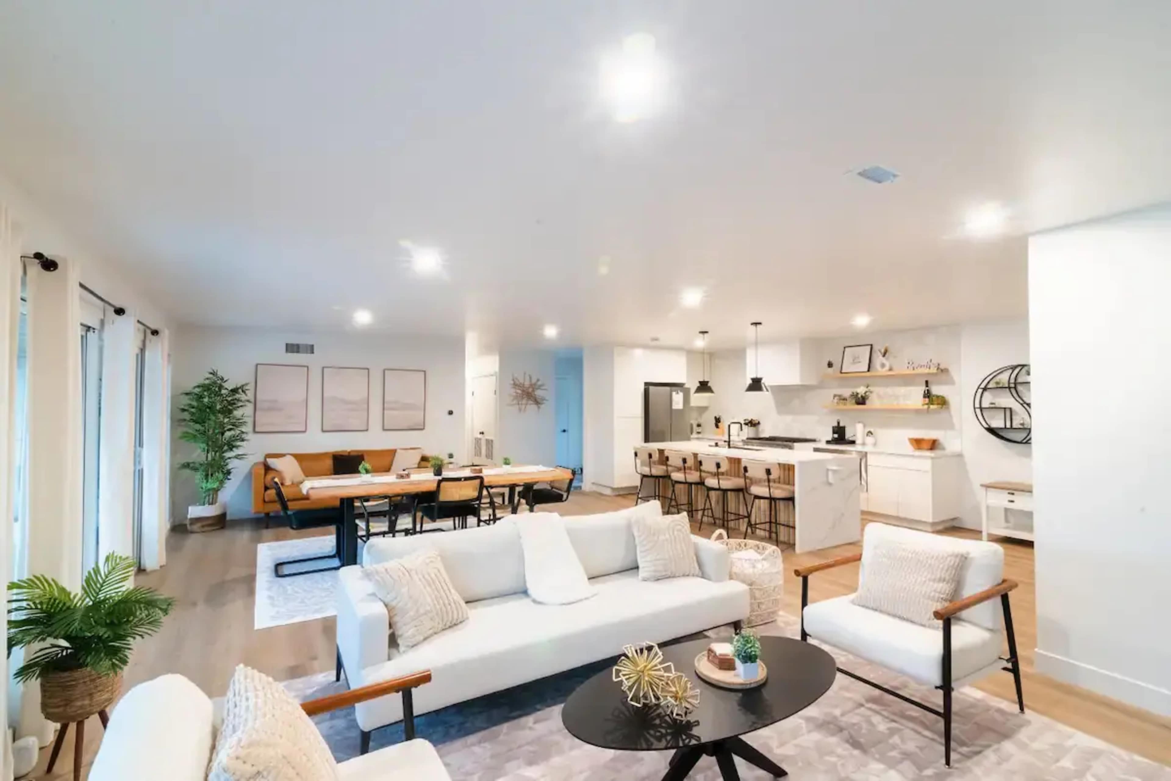 A modern open-concept living room and kitchen with a light-colored sofa, a central coffee table, a dining table, and bar seating.