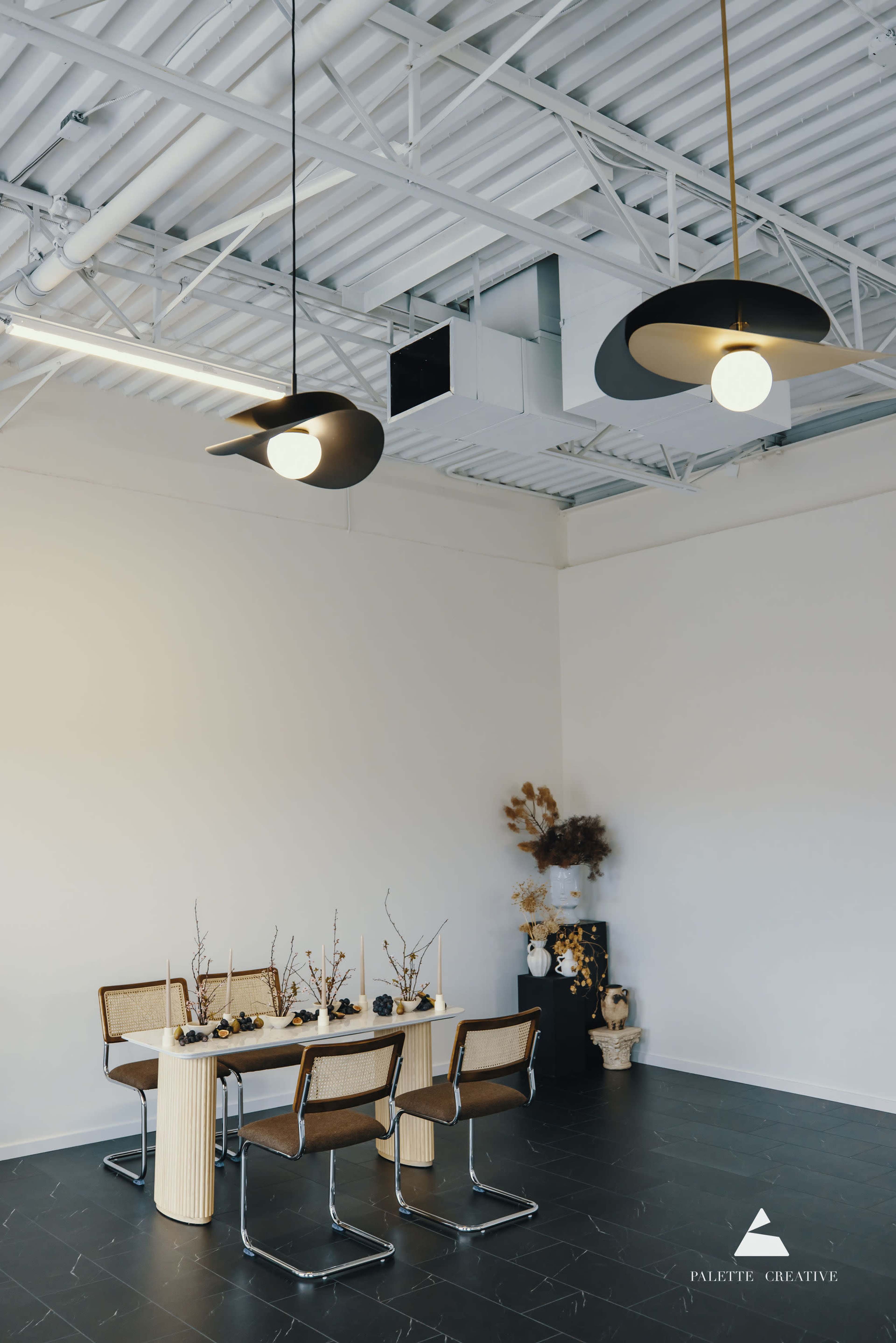The image shows a minimalist dining area featuring a wooden table surrounded by chairs, with modern pendant lights and decorative elements along the wall.