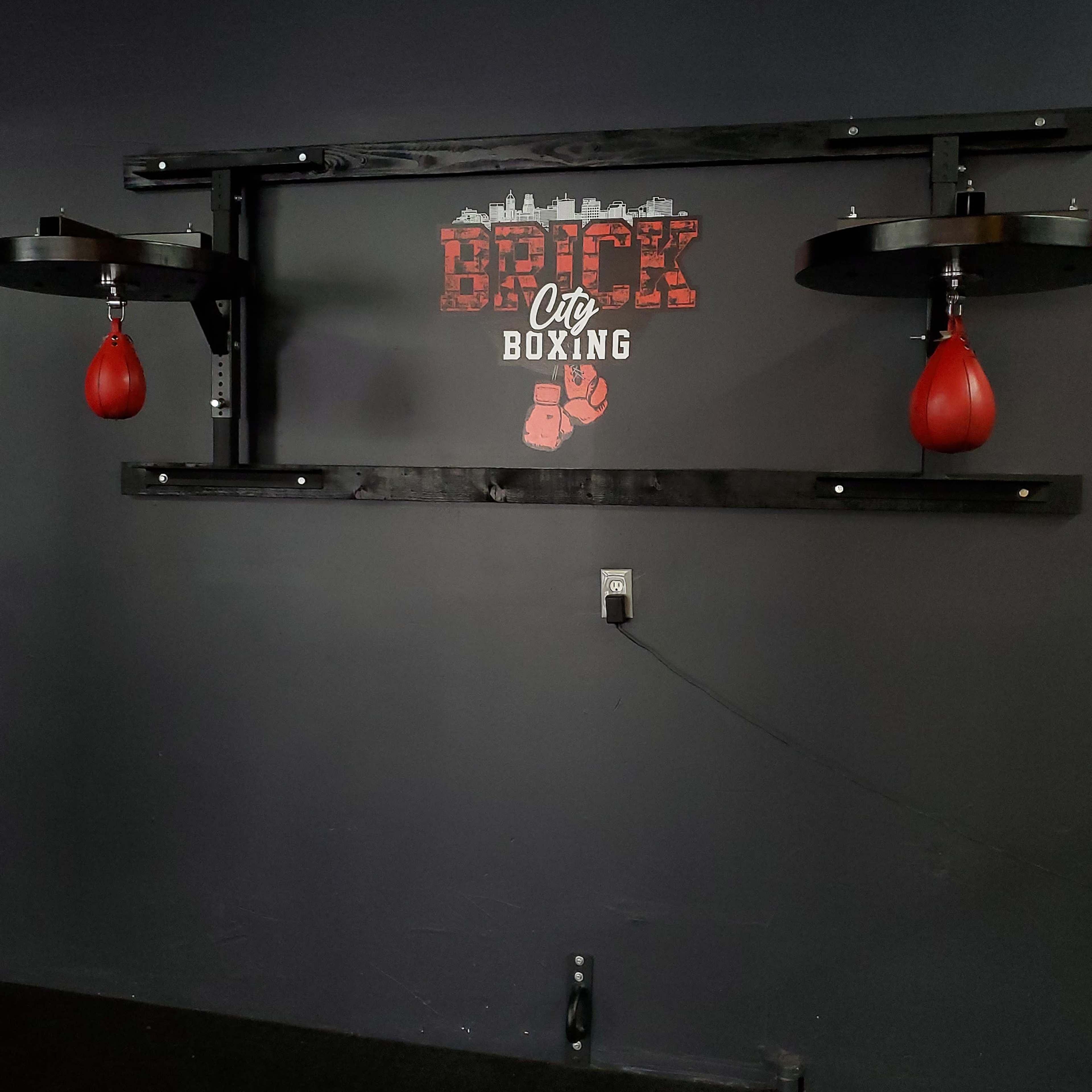 A boxing gym wall adorned with a large "Brick City Boxing" sign and two red punching bags mounted on a black wooden frame.