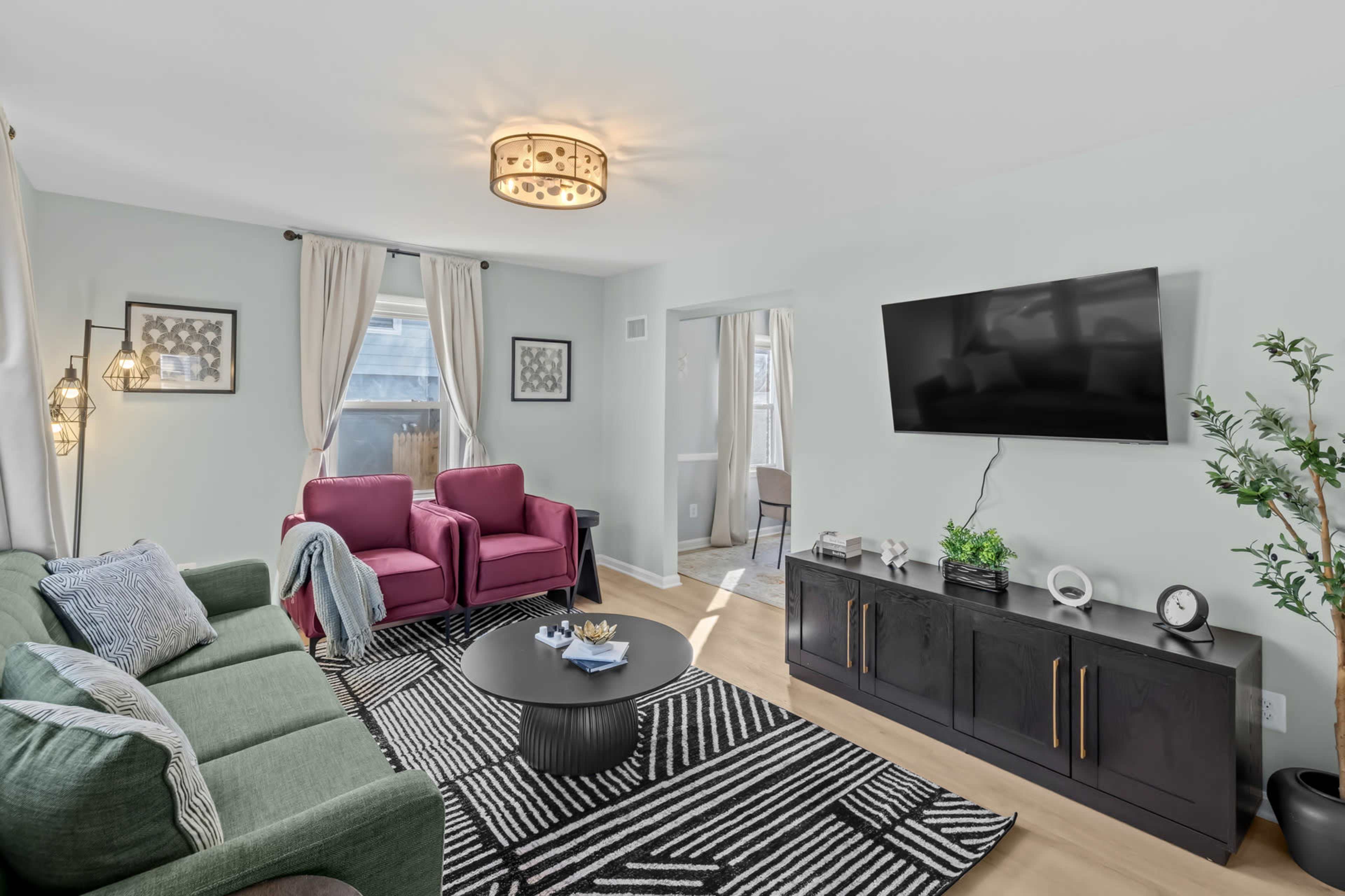 The image shows a modern living room featuring a green sofa, two red armchairs, a black coffee table, and a wall-mounted television, with large windows allowing natural light to enter.