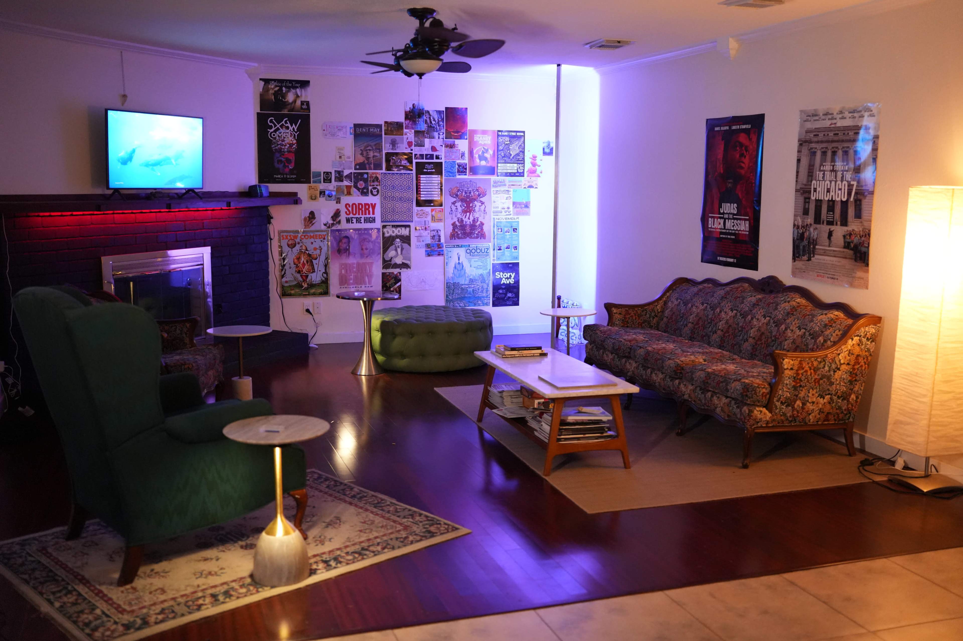 The room features a combination of vintage furniture, including a floral sofa and a green armchair, with walls adorned by various posters and a television mounted in the corner.