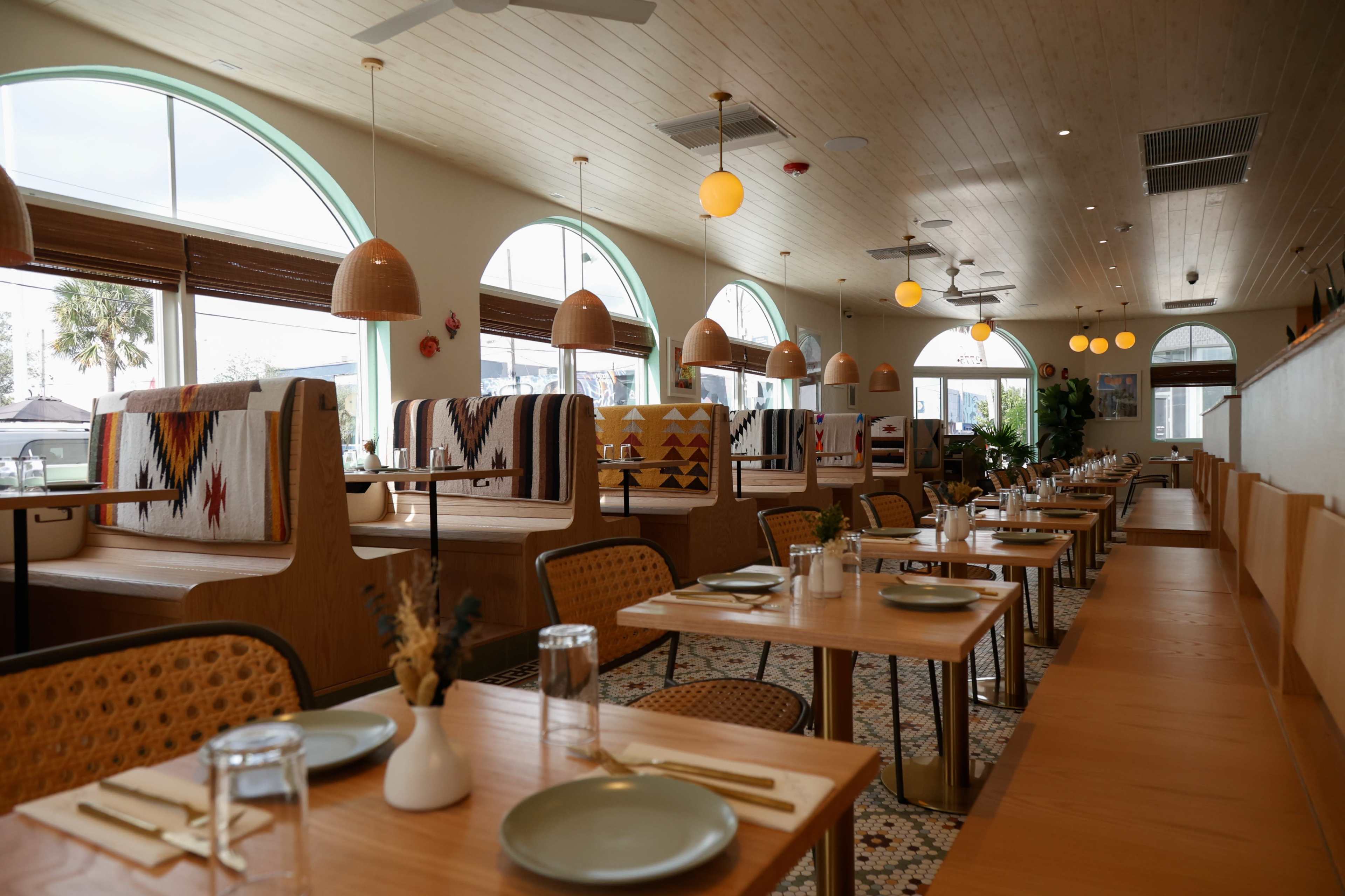 The image shows an interior of a restaurant featuring wooden tables, cozy seating booths, and large arched windows allowing natural light to fill the space.