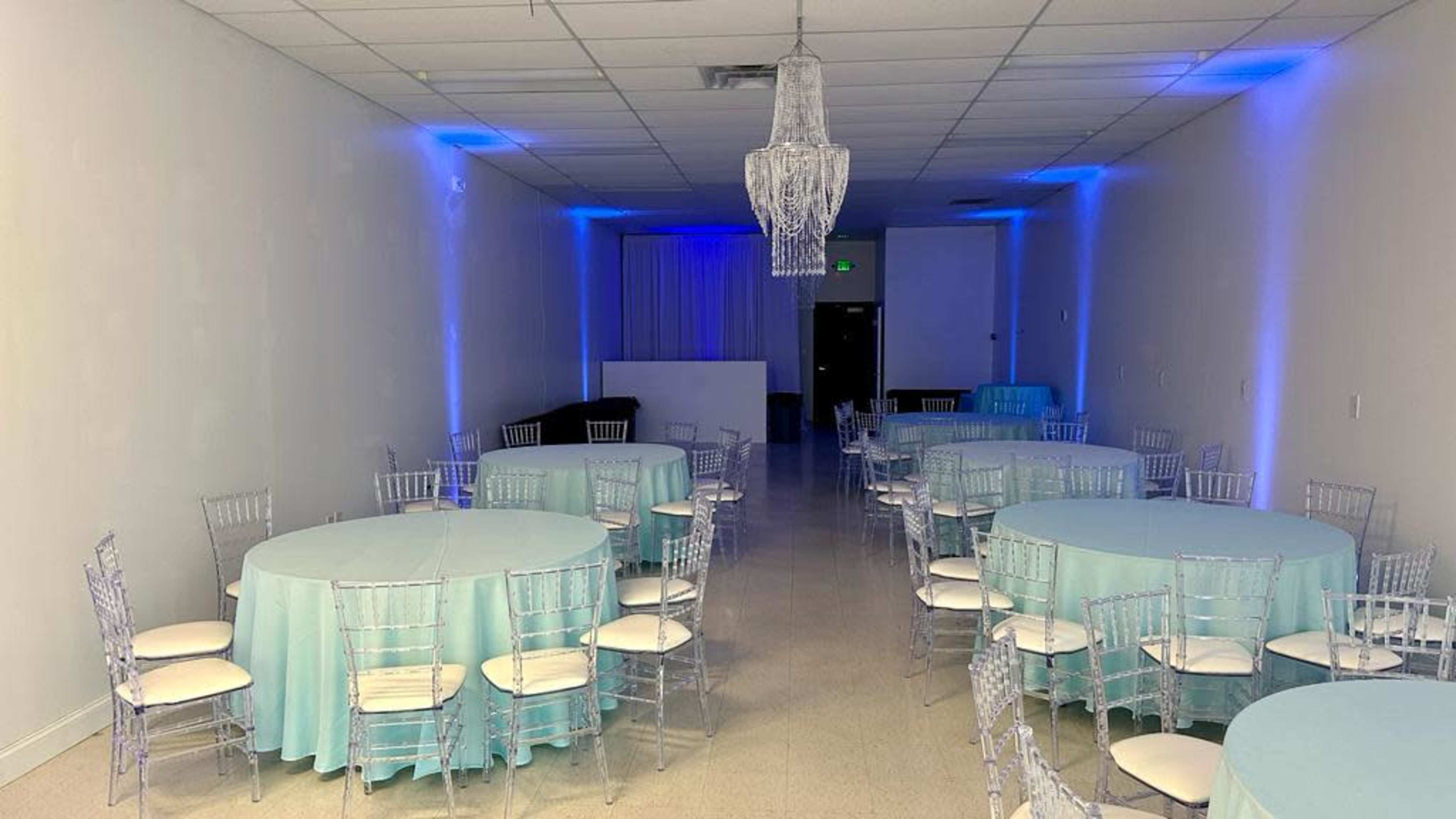 The image shows a spacious event room with round tables covered in light blue tablecloths and clear Chiavari chairs, illuminated by blue lighting and featuring a chandelier.