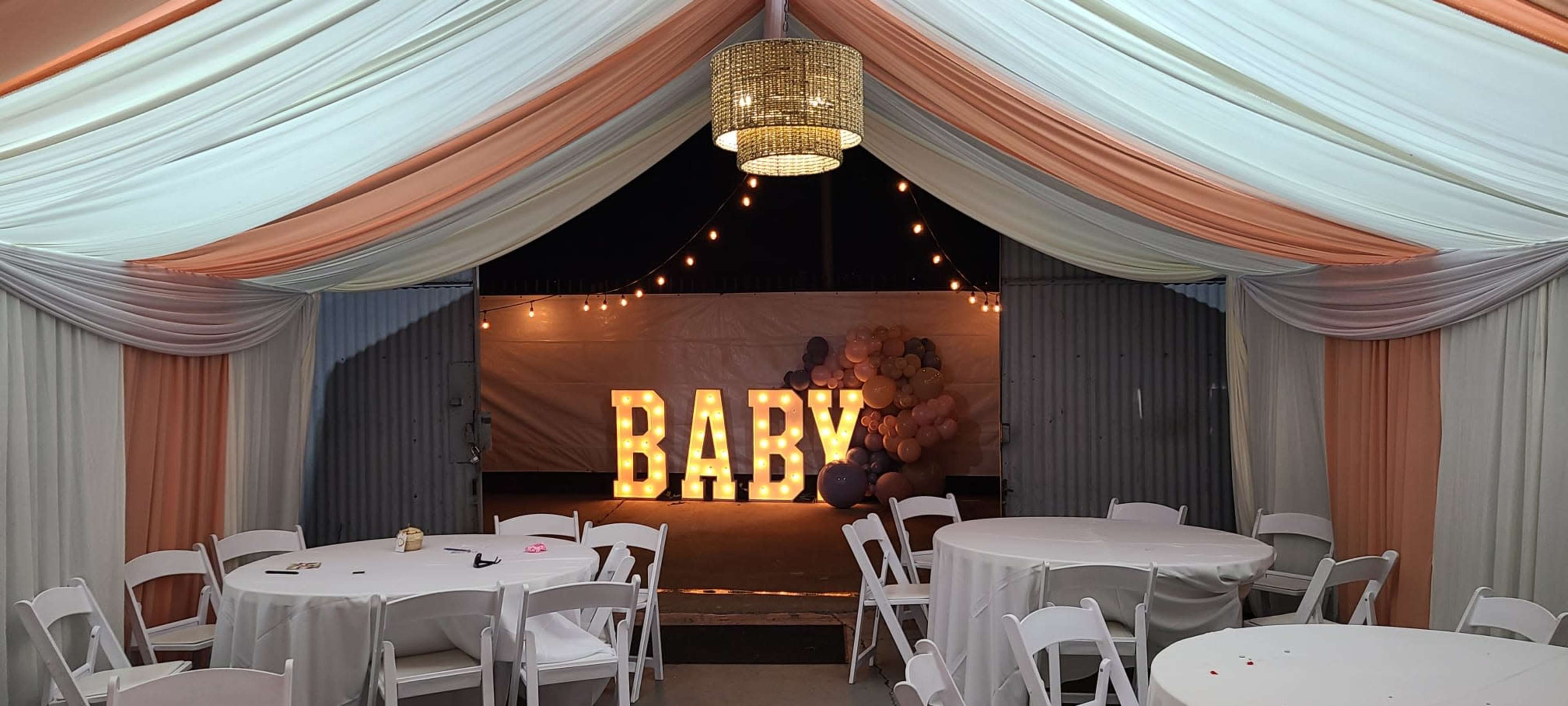 The scene shows a decorated indoor space featuring tables, draped fabric in orange and white, and a large illuminated "BABY" sign surrounded by balloons in the background.