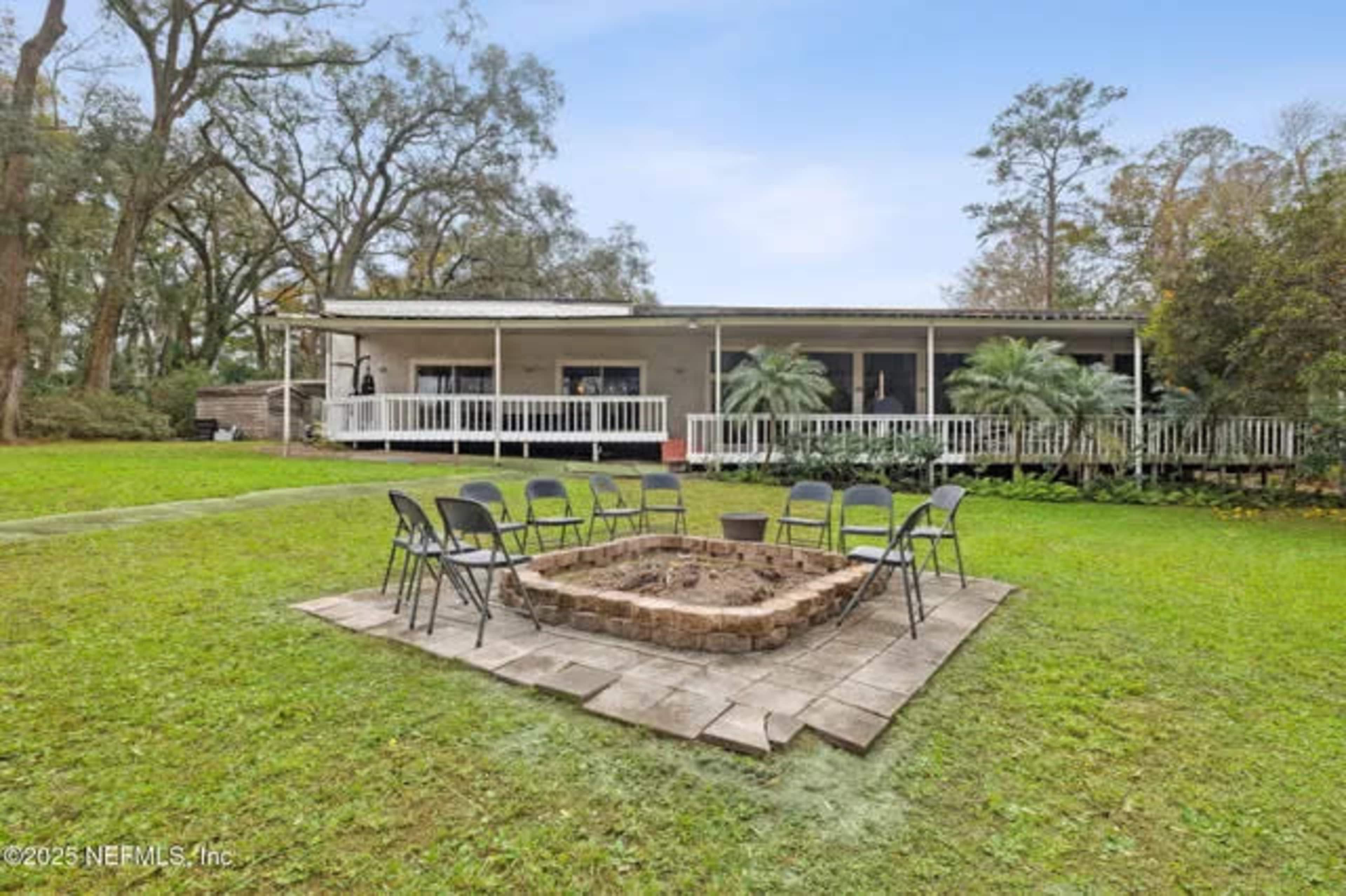 A circular stone fire pit surrounded by folding chairs is located in a grassy area in front of a single-story house with a porch.