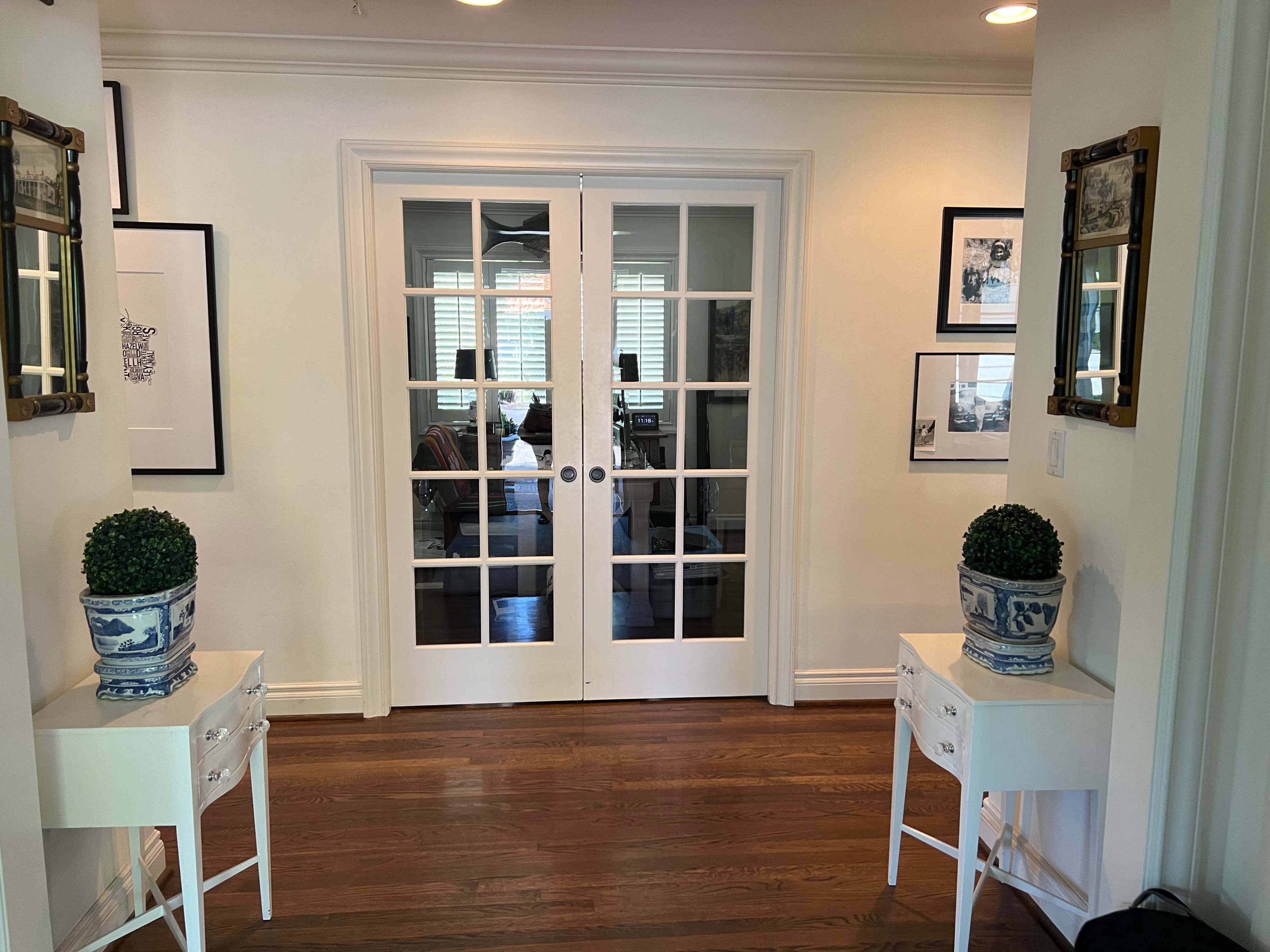 A hallway with two white side tables and potted plants, leading to double glass doors that open into a room.