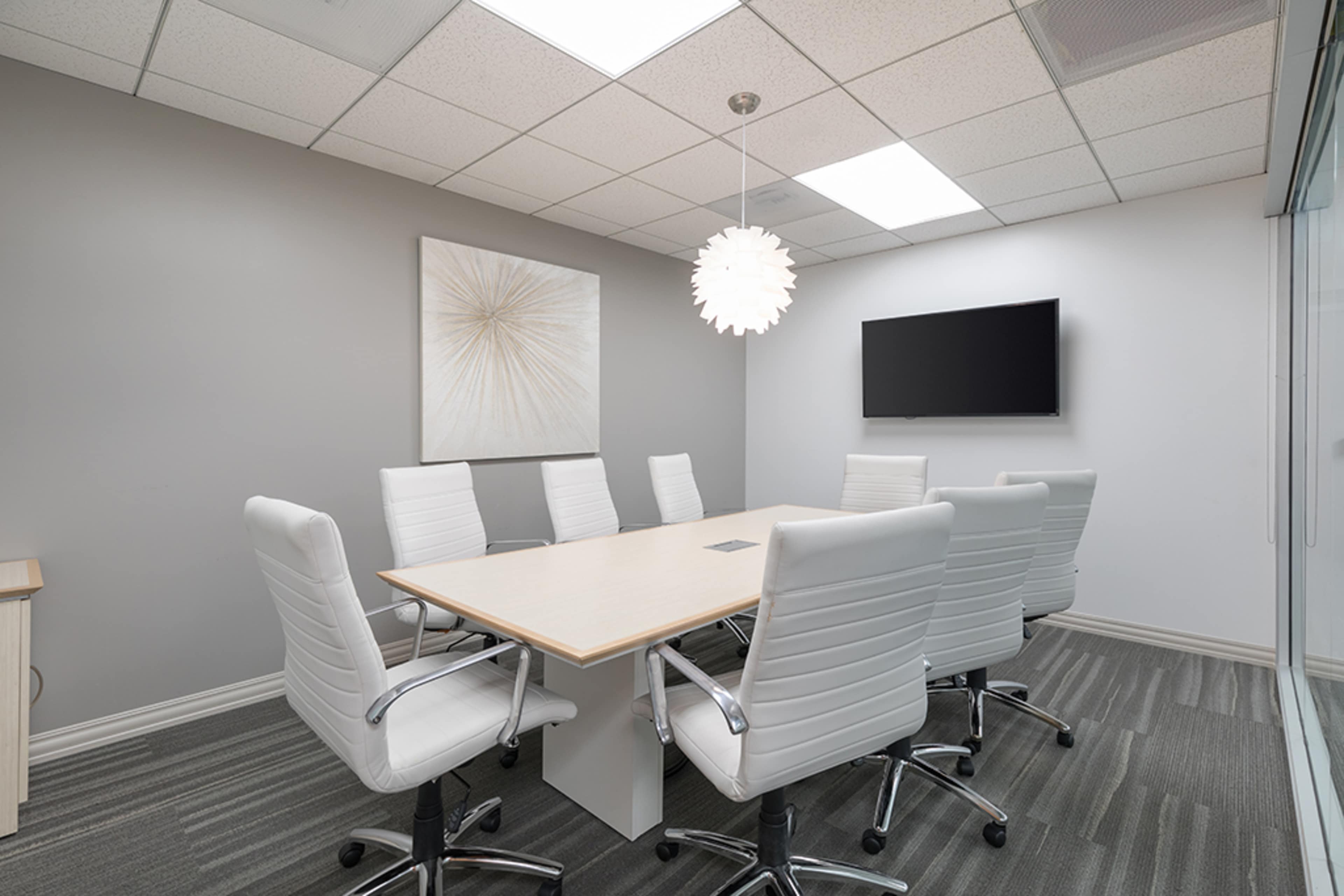 A modern conference room features a long table surrounded by six white chairs, a wall-mounted television, and abstract art hanging on the wall.