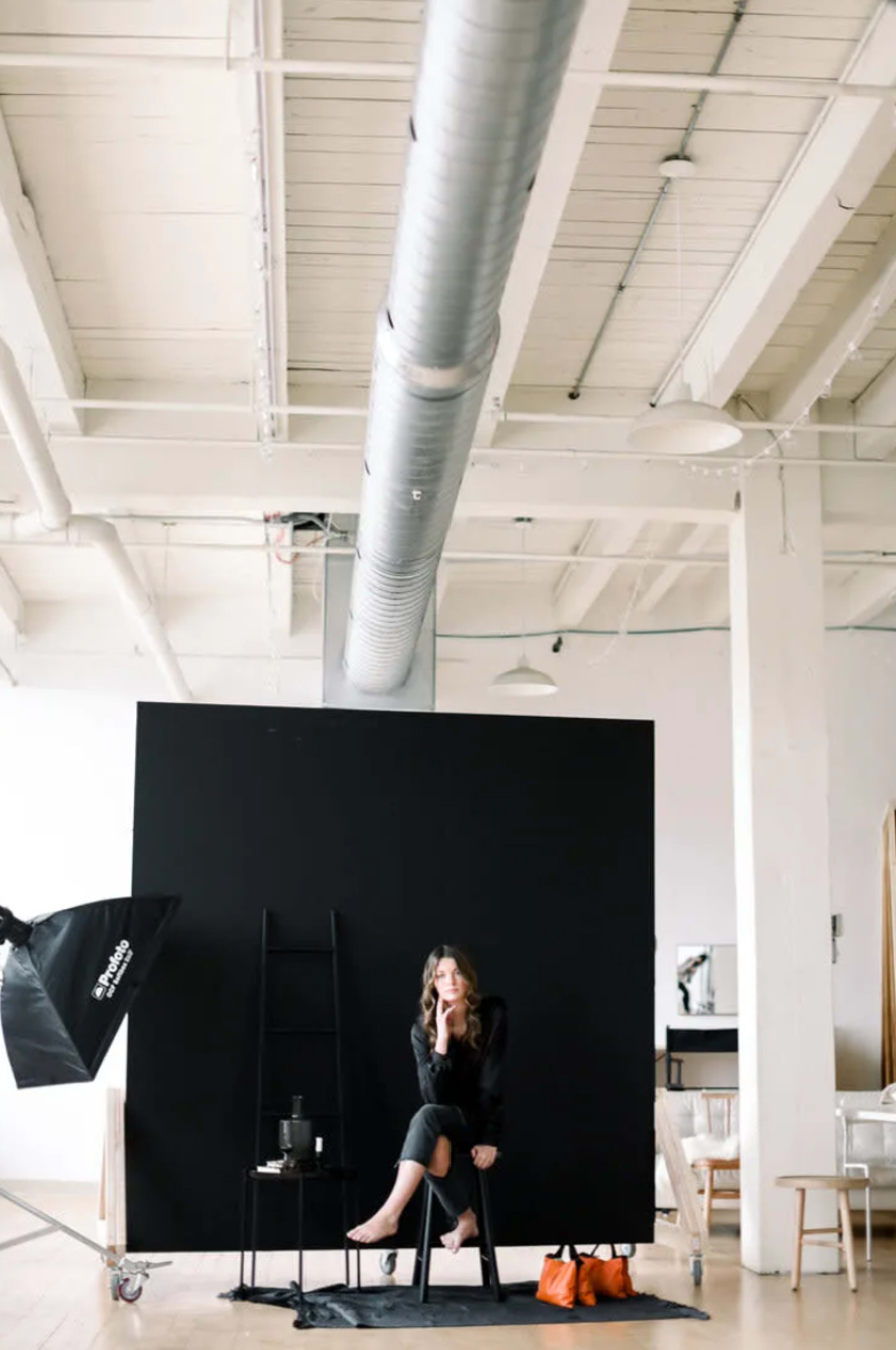 A woman sits on a stool in front of a black backdrop in a well-lit studio.