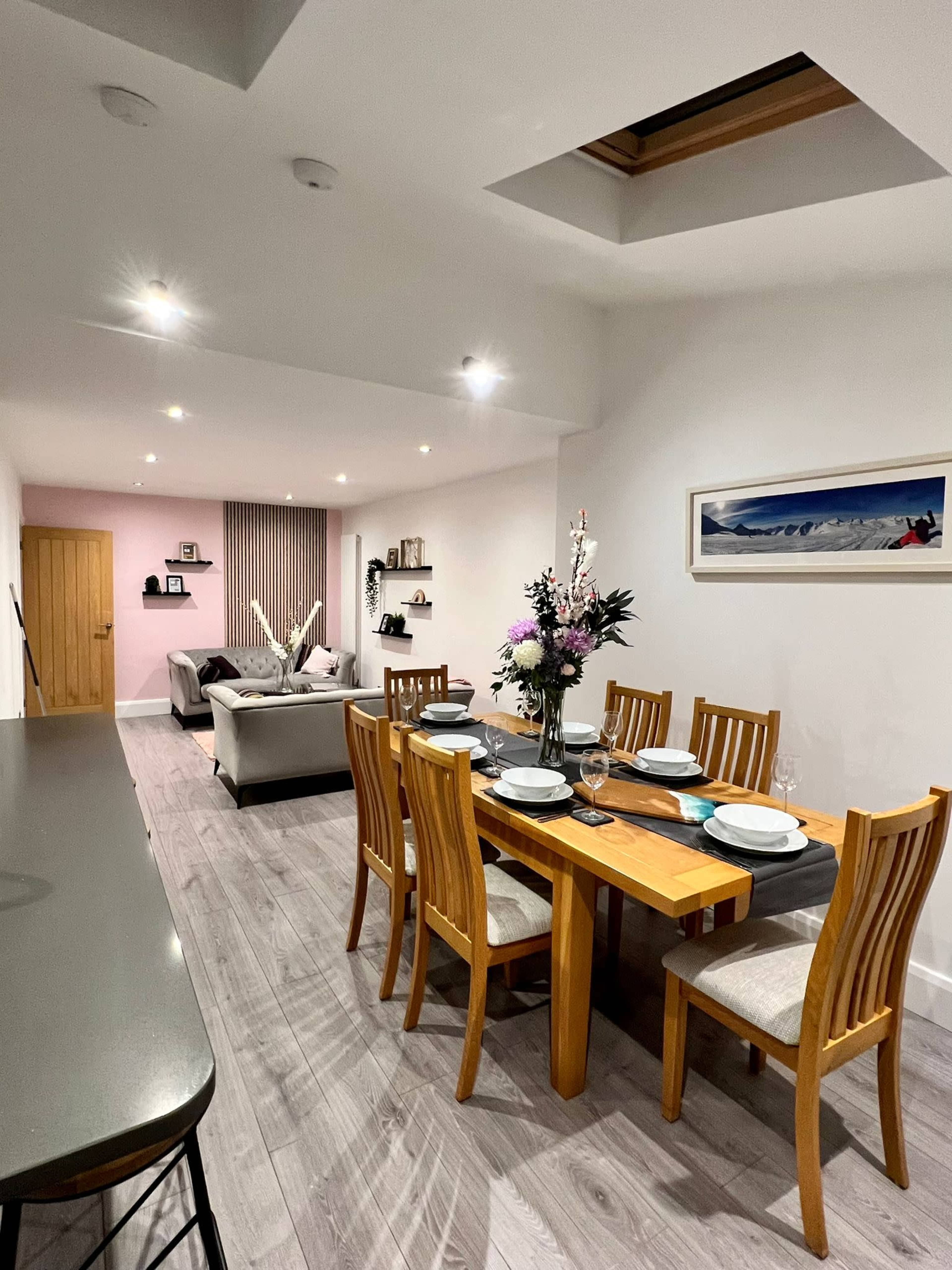 A dining area features a wooden table set for a meal, with a flower arrangement as the centerpiece, alongside a living space visible in the background.