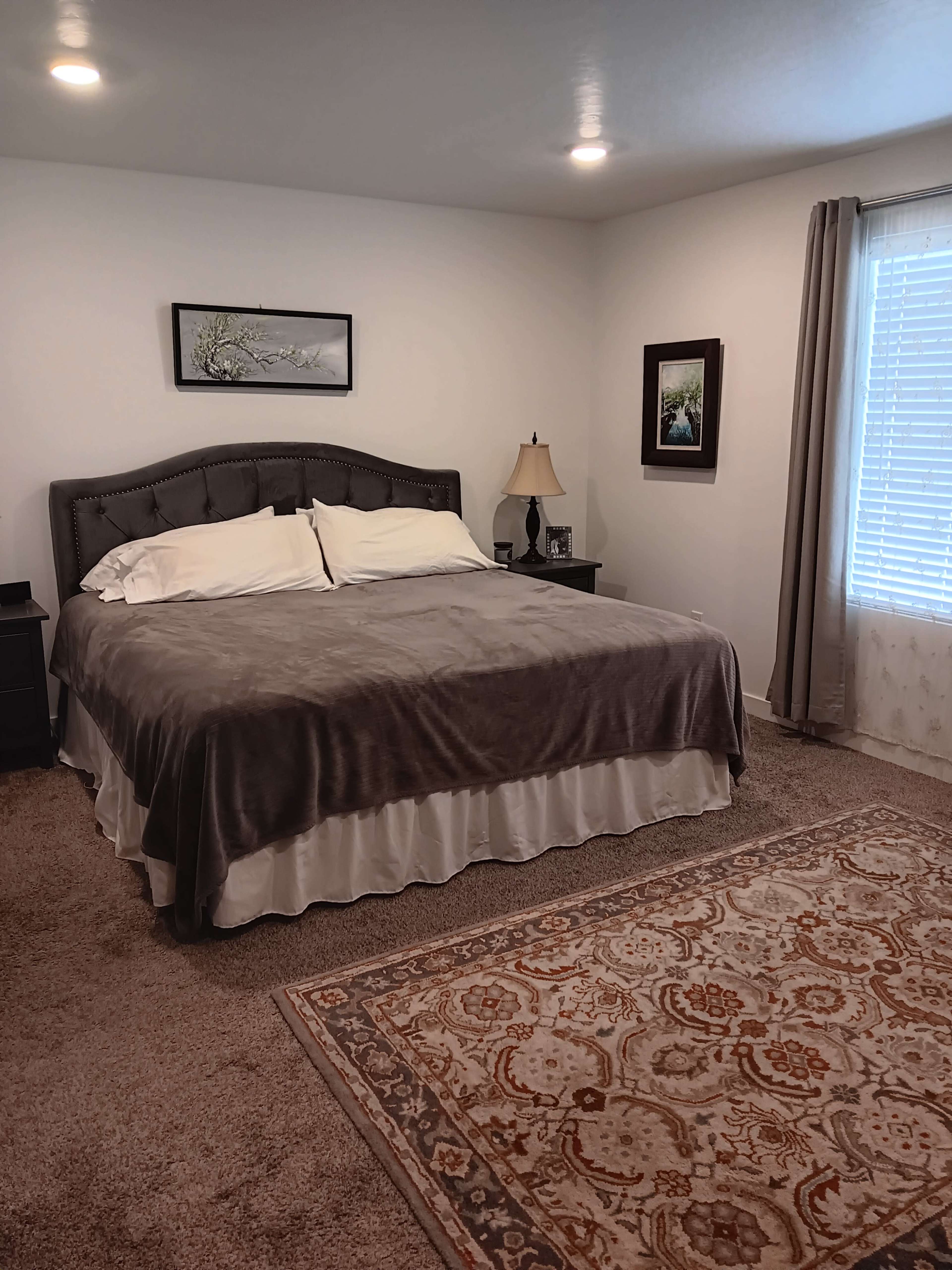 The image shows a neatly arranged bedroom with a large bed covered in a gray blanket, bedside tables with lamps, and a window with light-colored curtains.