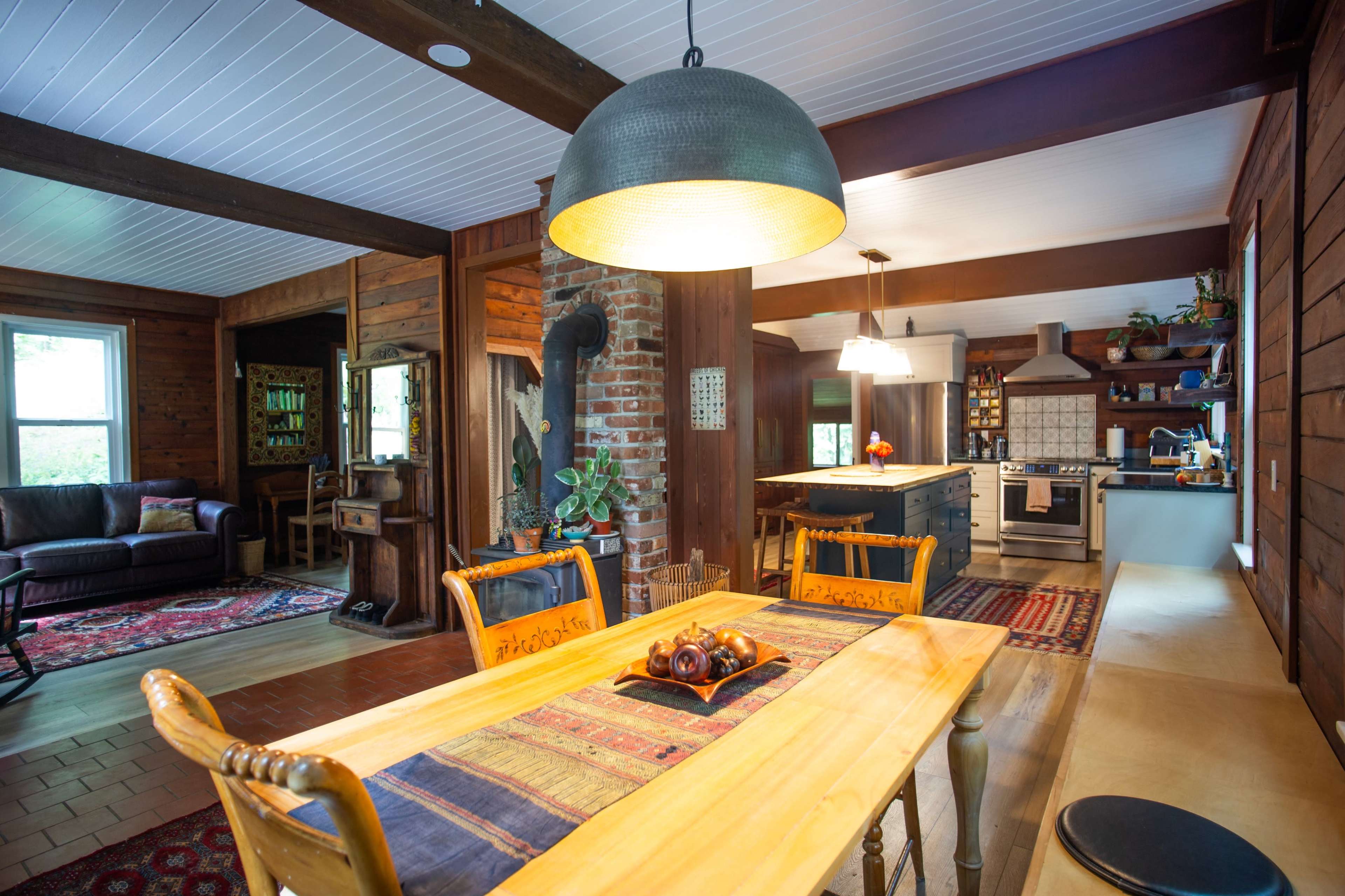 The image shows a wooden dining area with a table set for meals, adjacent to a kitchen featuring modern appliances and rustic decor.