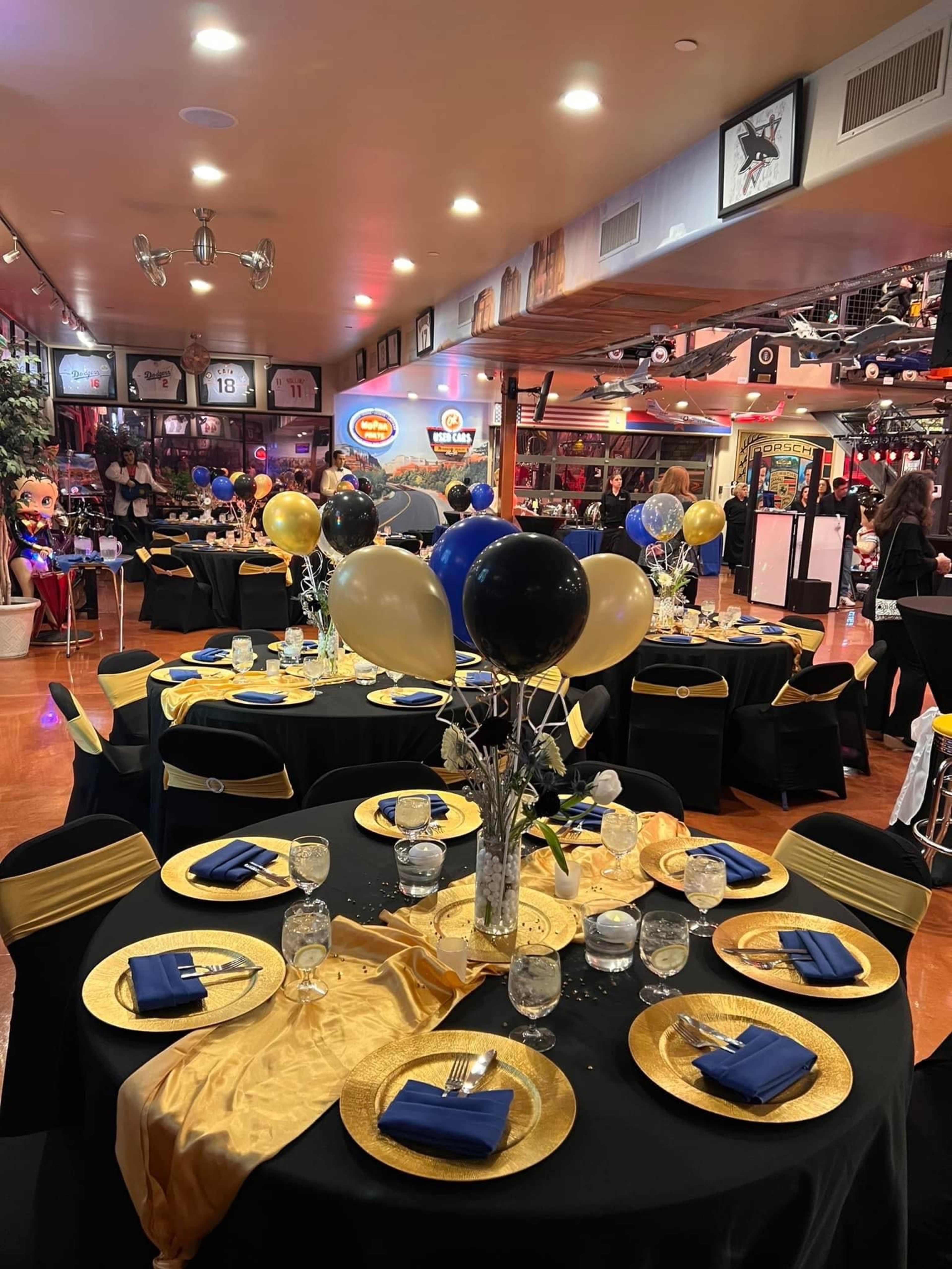 A banquet hall is set up with elegantly decorated tables featuring black and gold tablecloths, plates, and balloons.