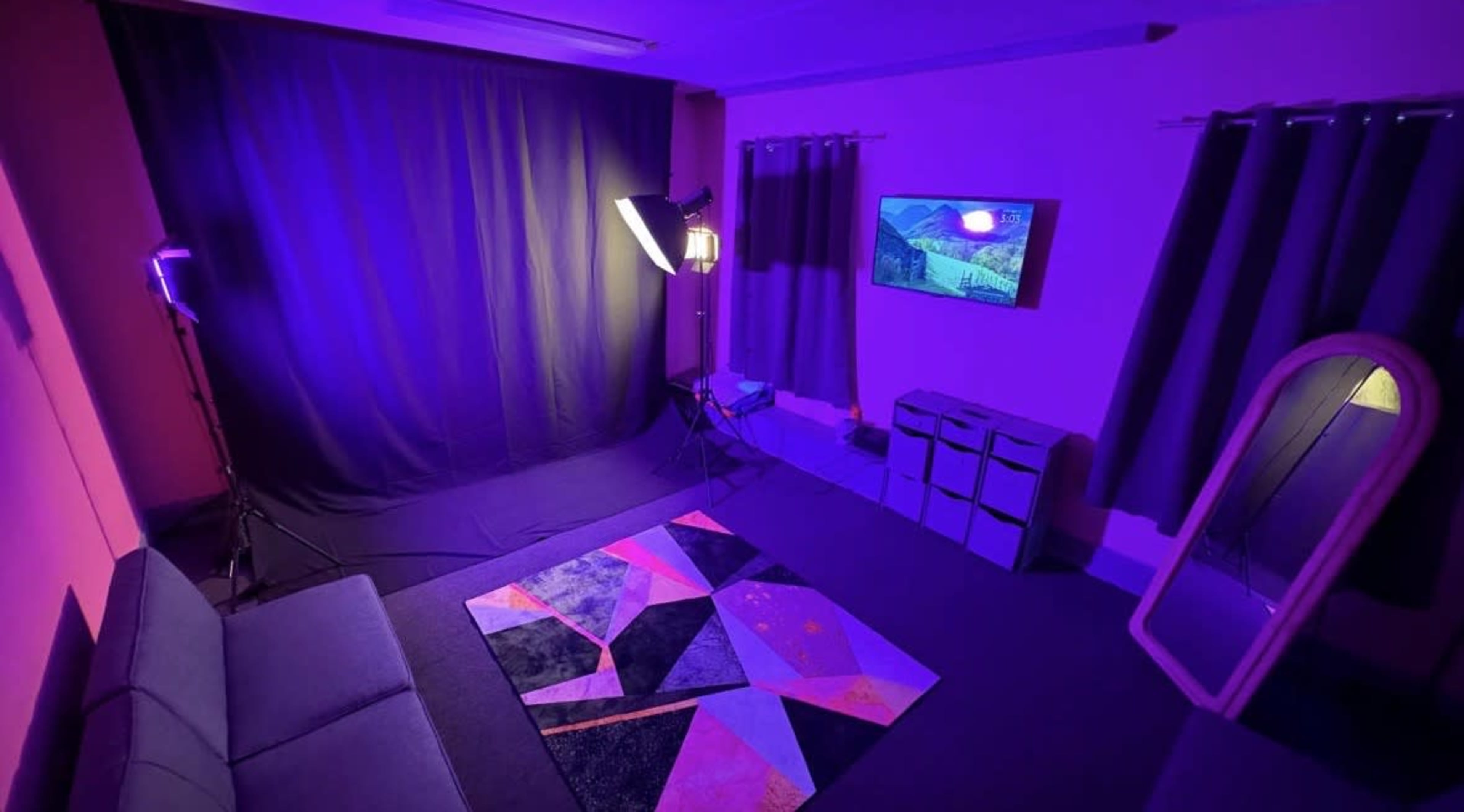 The image shows a dimly lit room with purple walls, a black backdrop, a softbox lighting setup, a large mirror, and a colorful geometric rug.