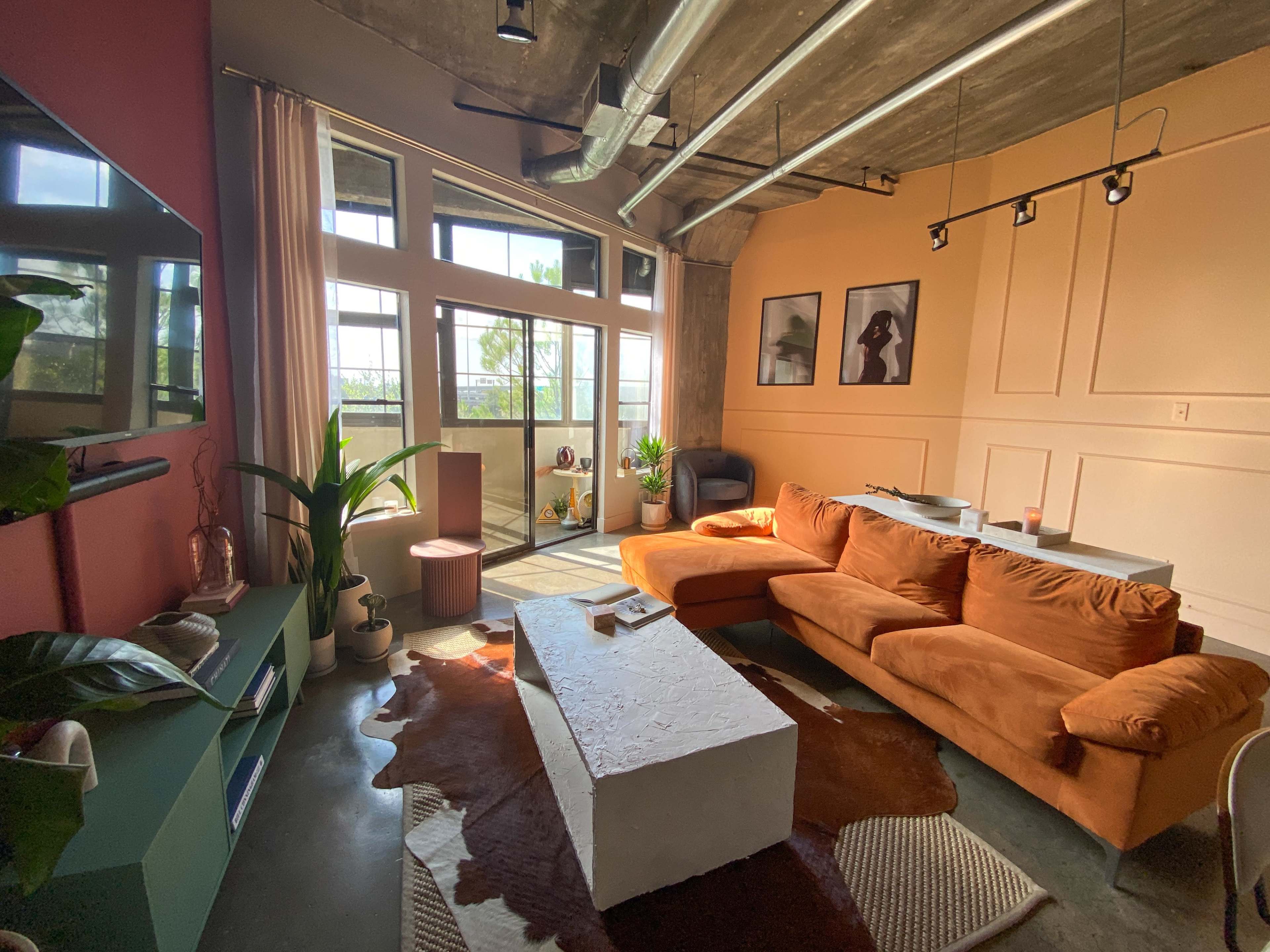 A modern living room with a large orange sectional sofa, a white coffee table, plants, and large windows that let in natural light.