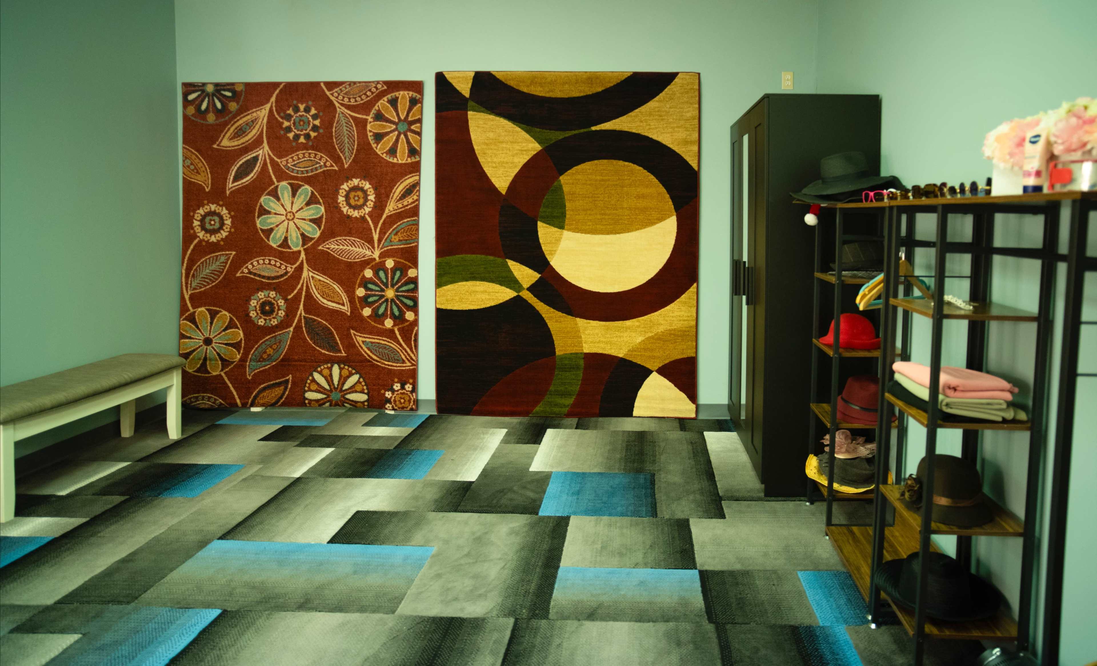 The image shows a room with two large area rugs on the wall and a storage unit filled with hats and accessories.