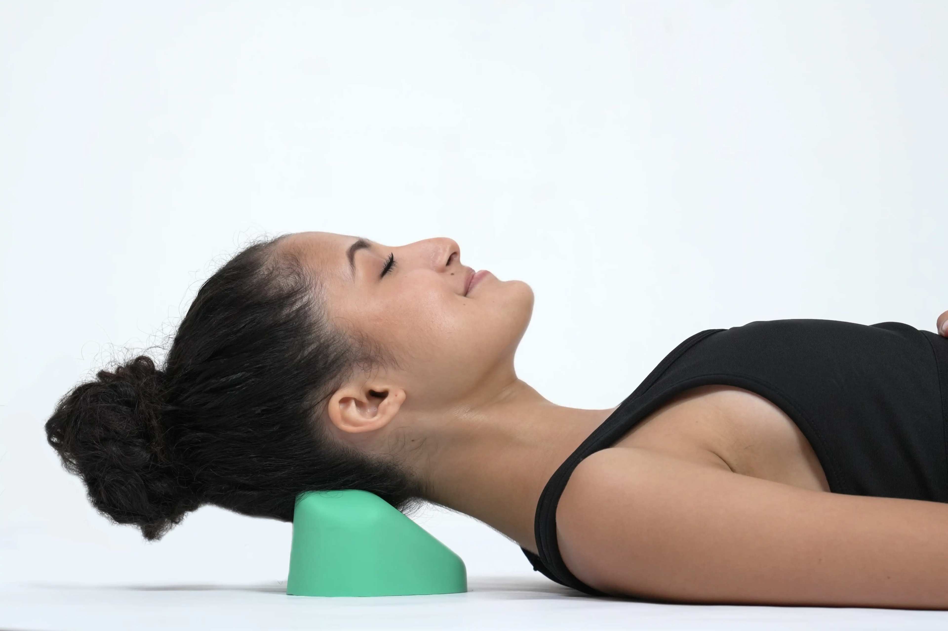 A woman lies on her back with her head supported by a green cushion on a white surface.