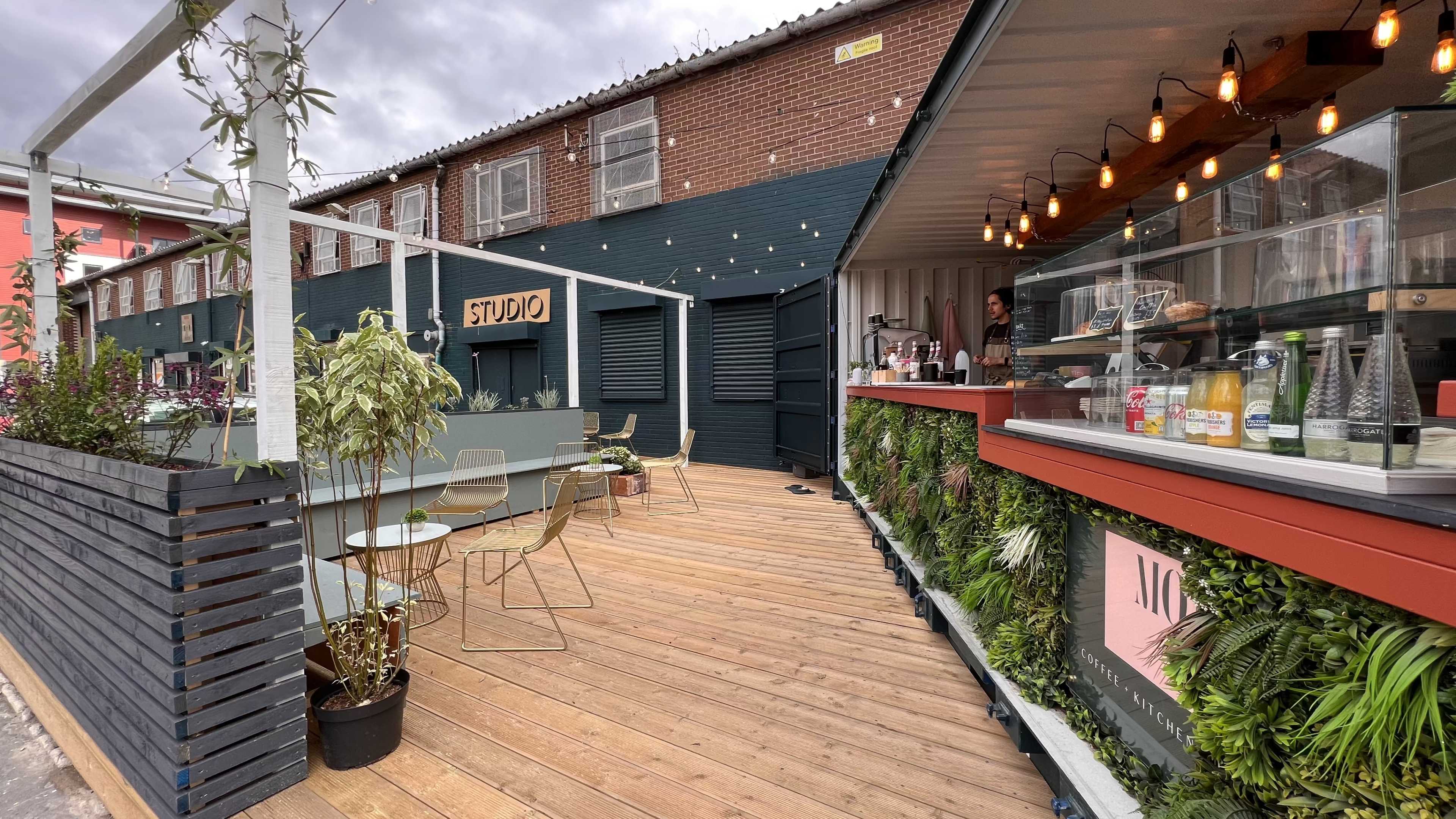 An outdoor café area with wooden decking, featuring plants, seating, and a service counter with drinks and food on display.