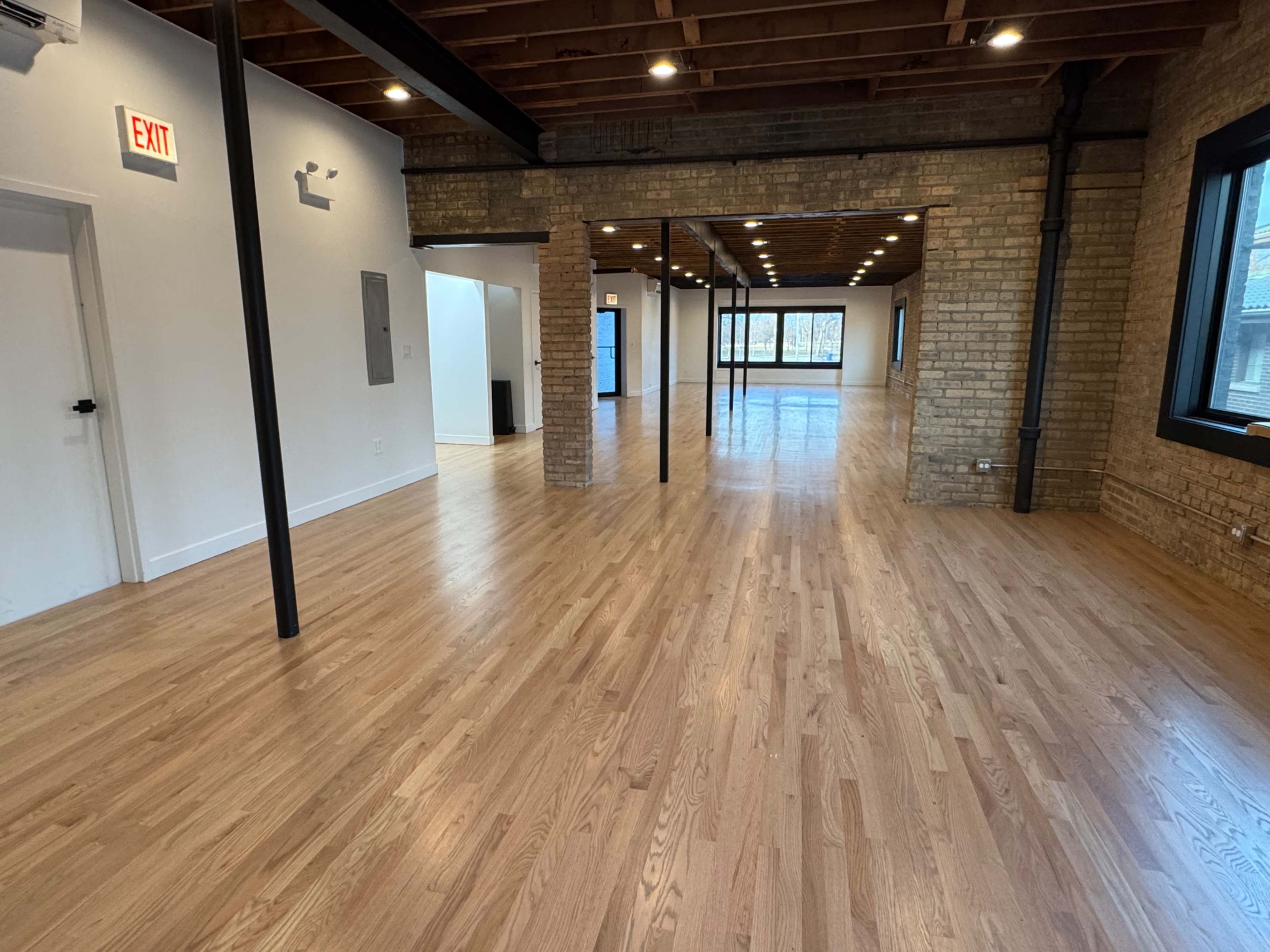 The image shows a spacious, empty interior with polished hardwood floors, exposed wooden beams, and large windows.