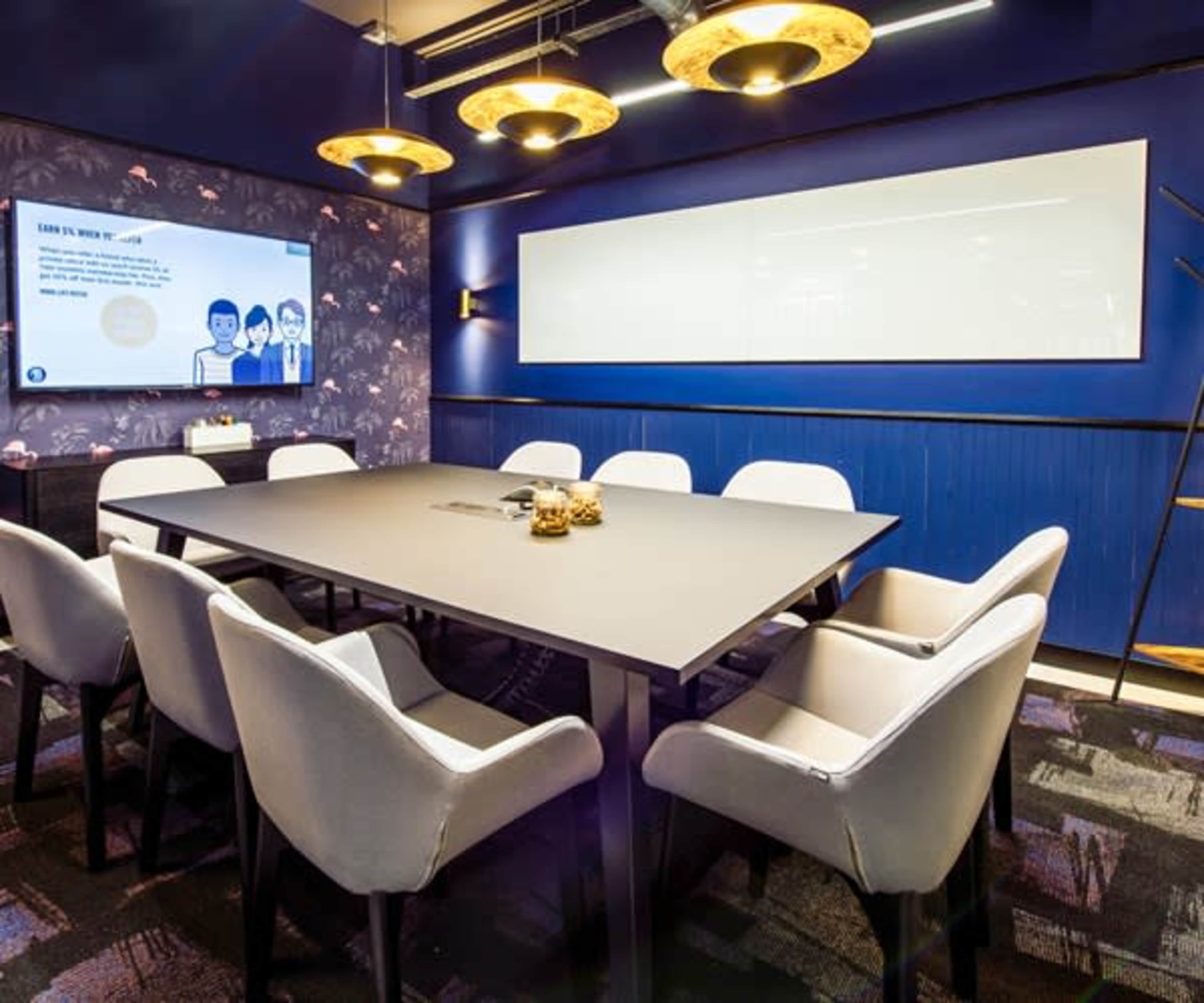 A conference room features a large rectangular table surrounded by upholstered chairs, with a projector screen and decorative lighting on the walls.