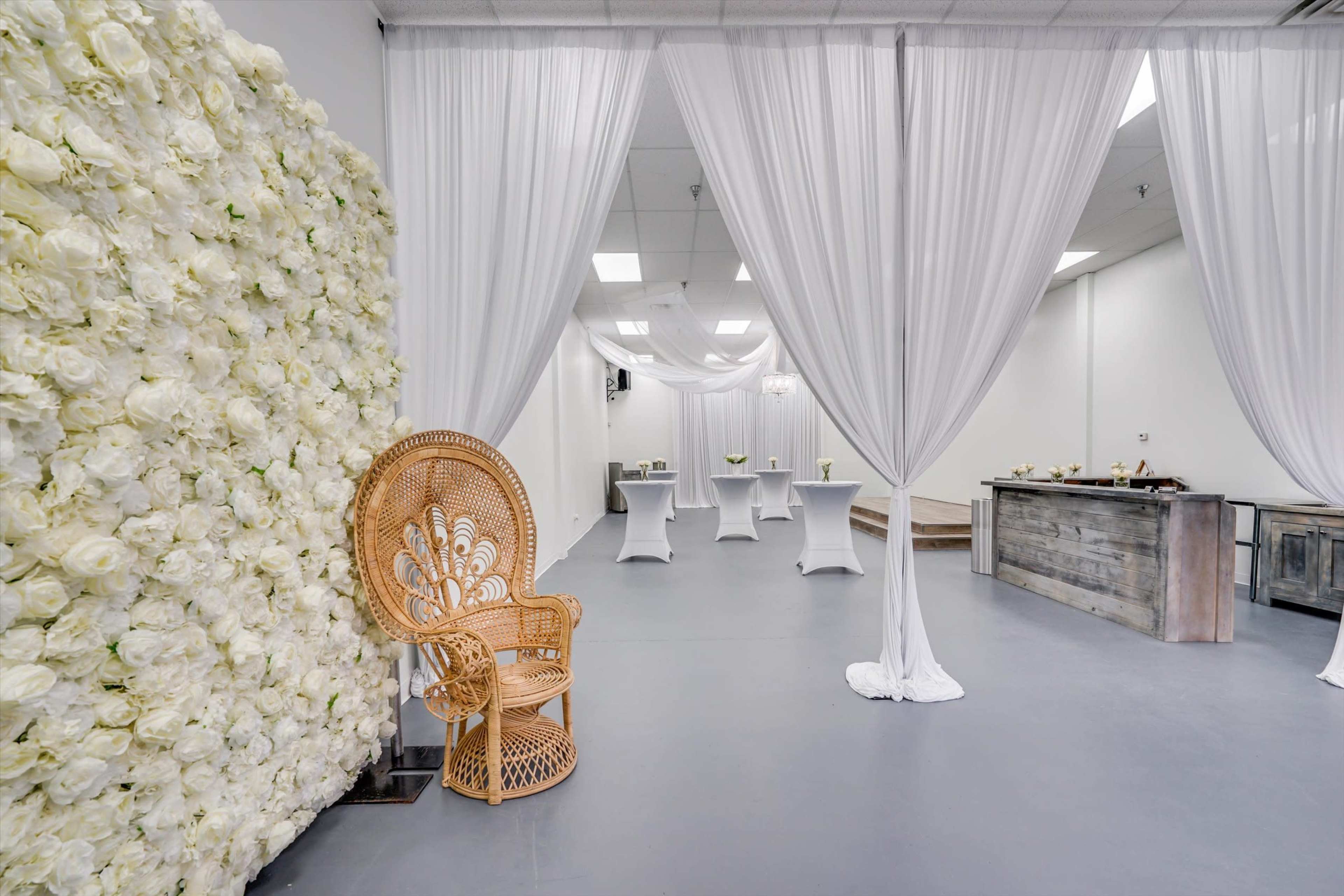 The image shows a spacious event room featuring a white floral wall, draped fabric, a rattan chair, and several tables set up for an occasion.