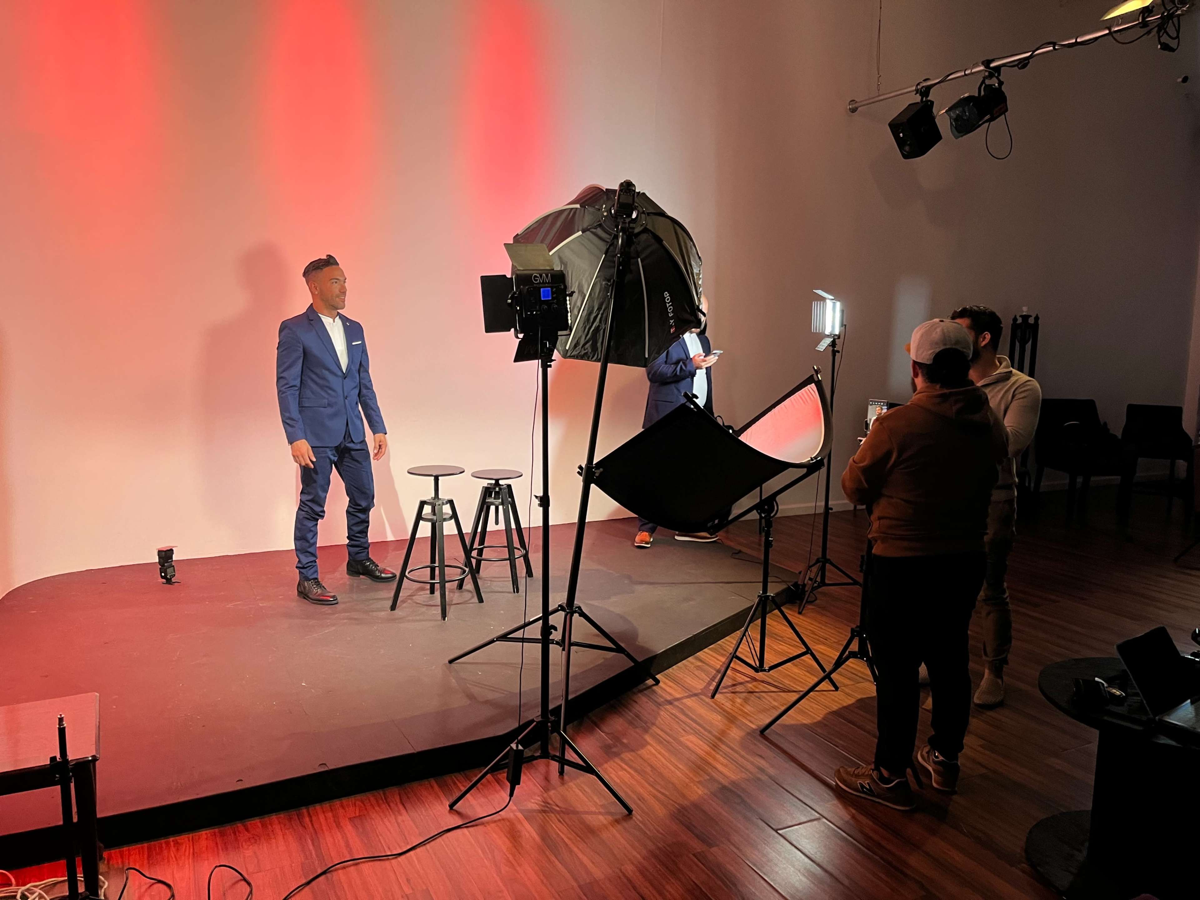 A man in a blue suit stands on a stage with two black stools, while a crew sets up lighting equipment in the background.