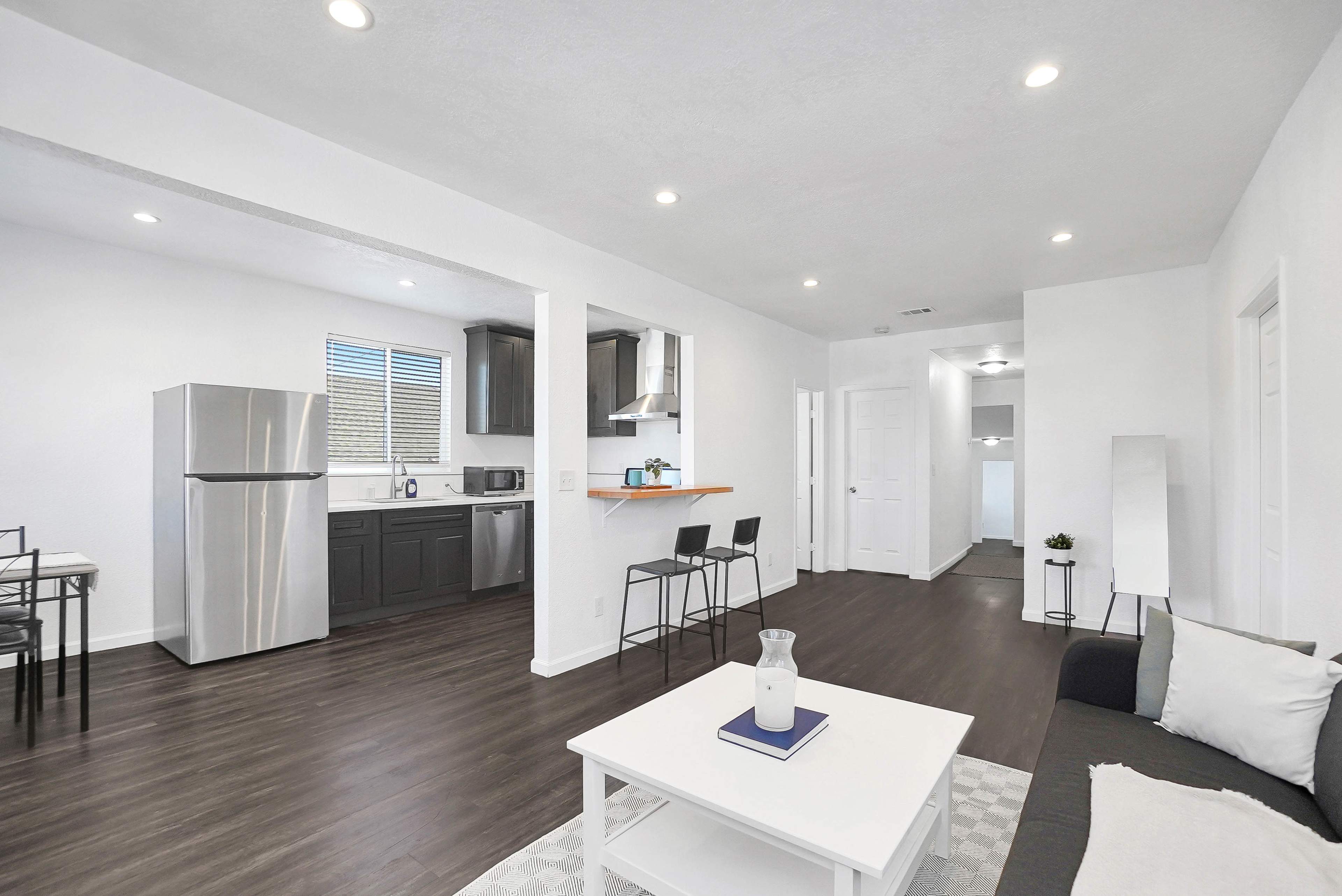 The image shows a modern, open-concept living space with a kitchen, dining area, and living room, featuring stainless steel appliances and minimalist furnishings.