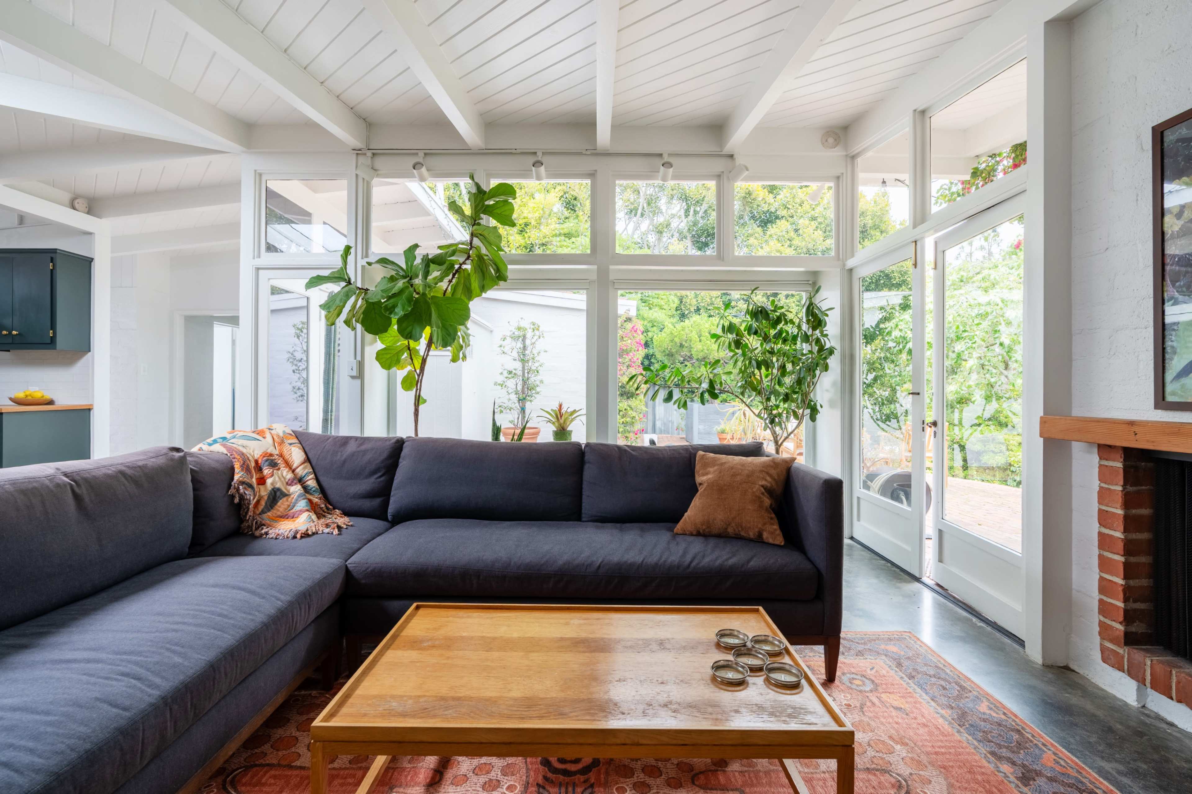 A modern living room features a large sectional sofa, a wooden coffee table, and numerous plants, with floor-to-ceiling windows allowing natural light to fill the space.
